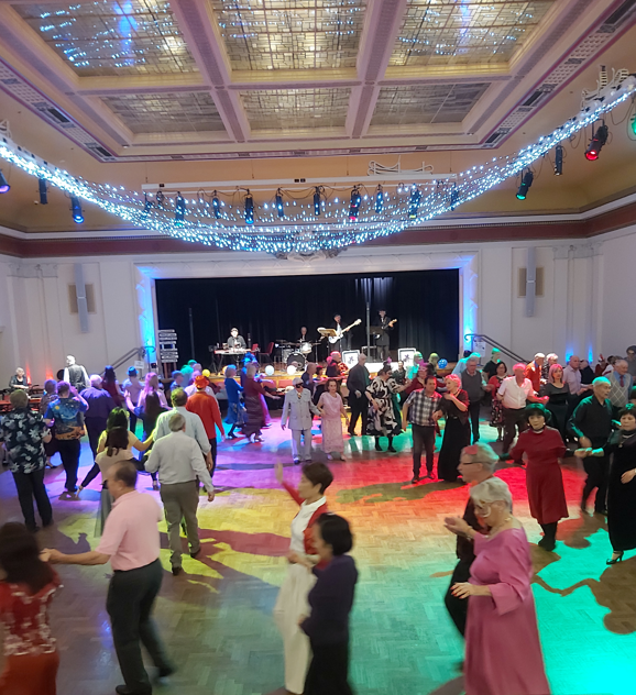 People dance together on a crowded dance floor while a live band performs on stage at a community social dance event.