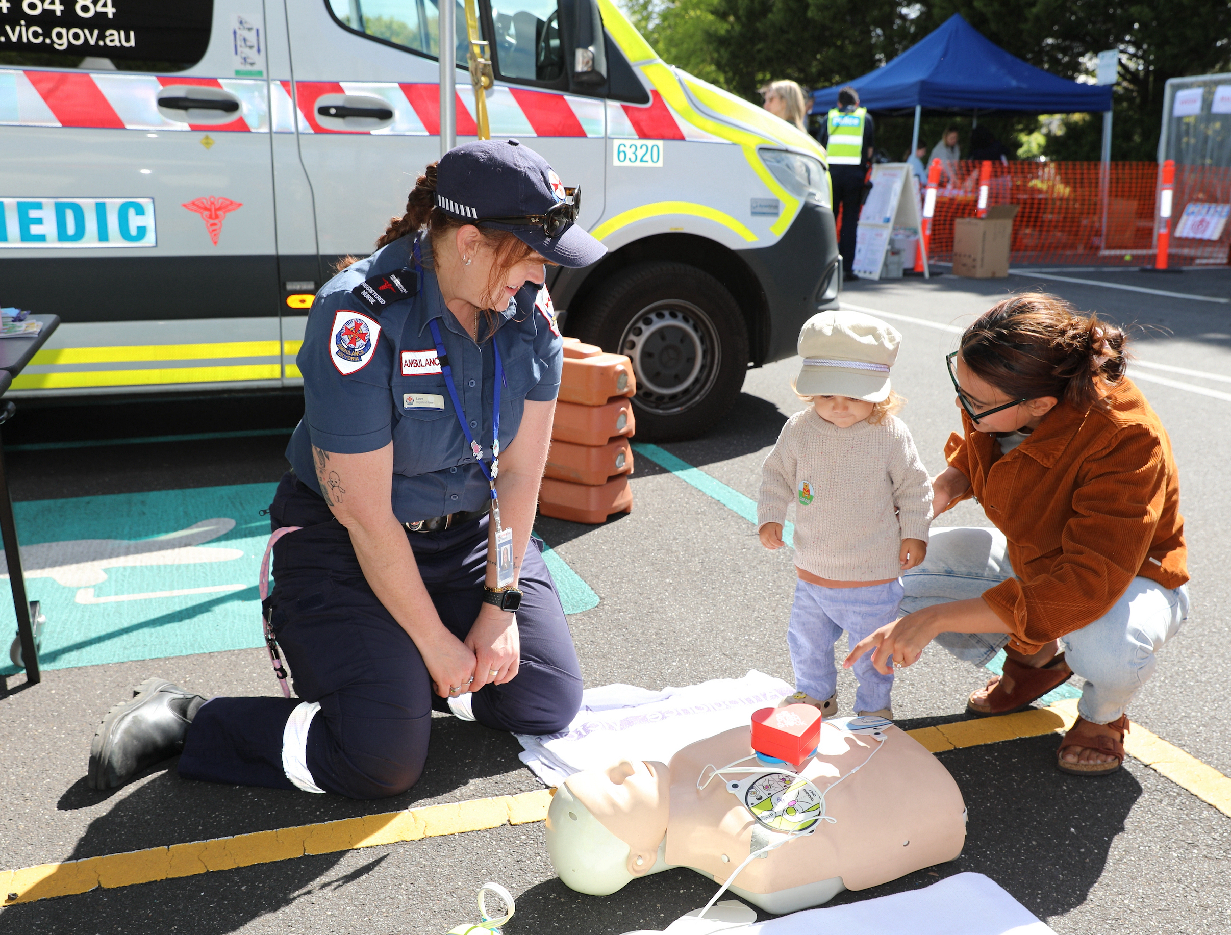 An Ambulance Victoria paramedic demonstrates CPR equipment to a child and caregiver at a community safety event, beside an ambulance.