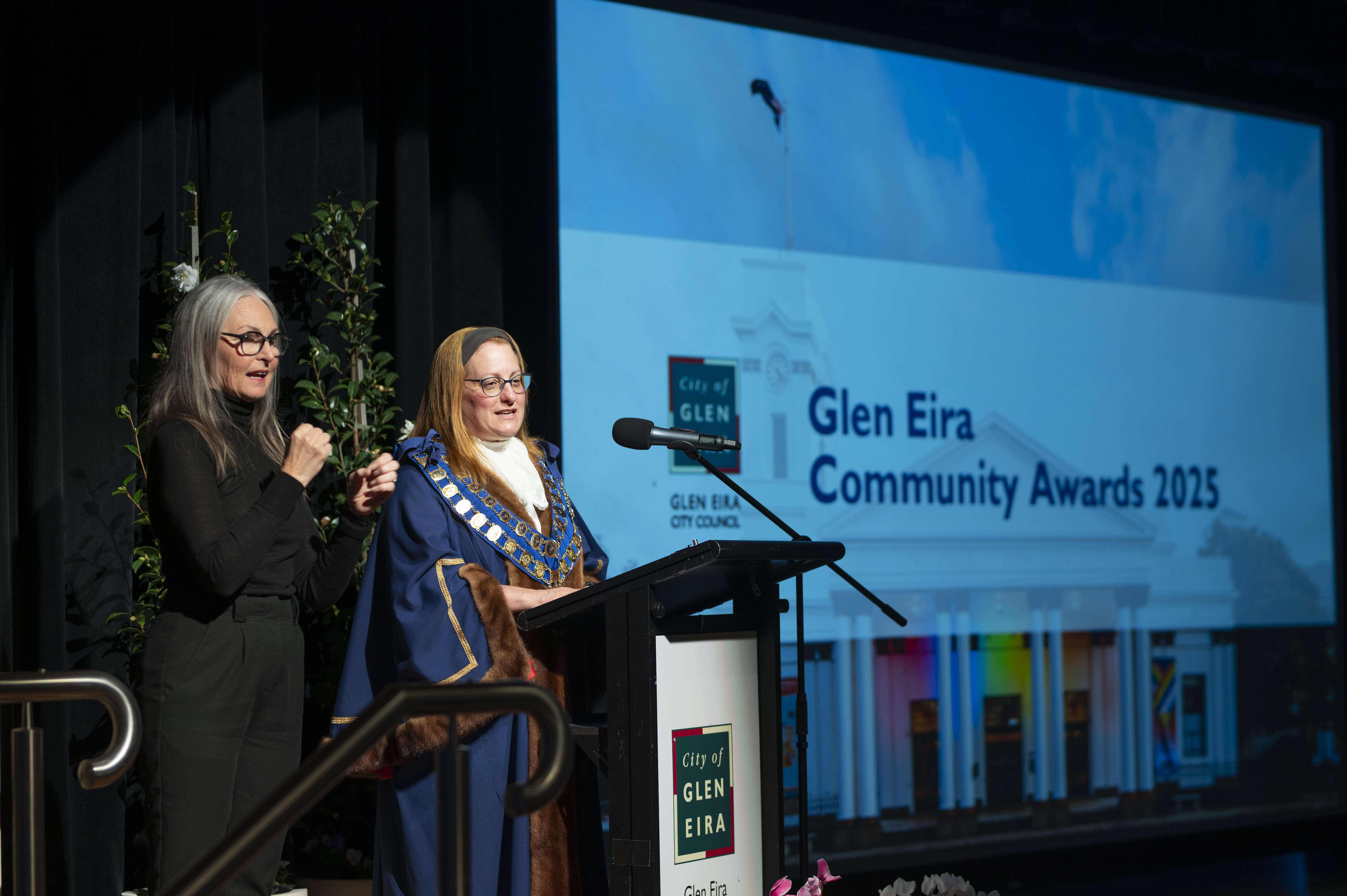 Two presenters standing at a podium during an event. One is dressed in ceremonial robes and regalia. Behind them, a large screen displays “Glen Eira Community Awards 2025” along with the City of Glen Eira logo.