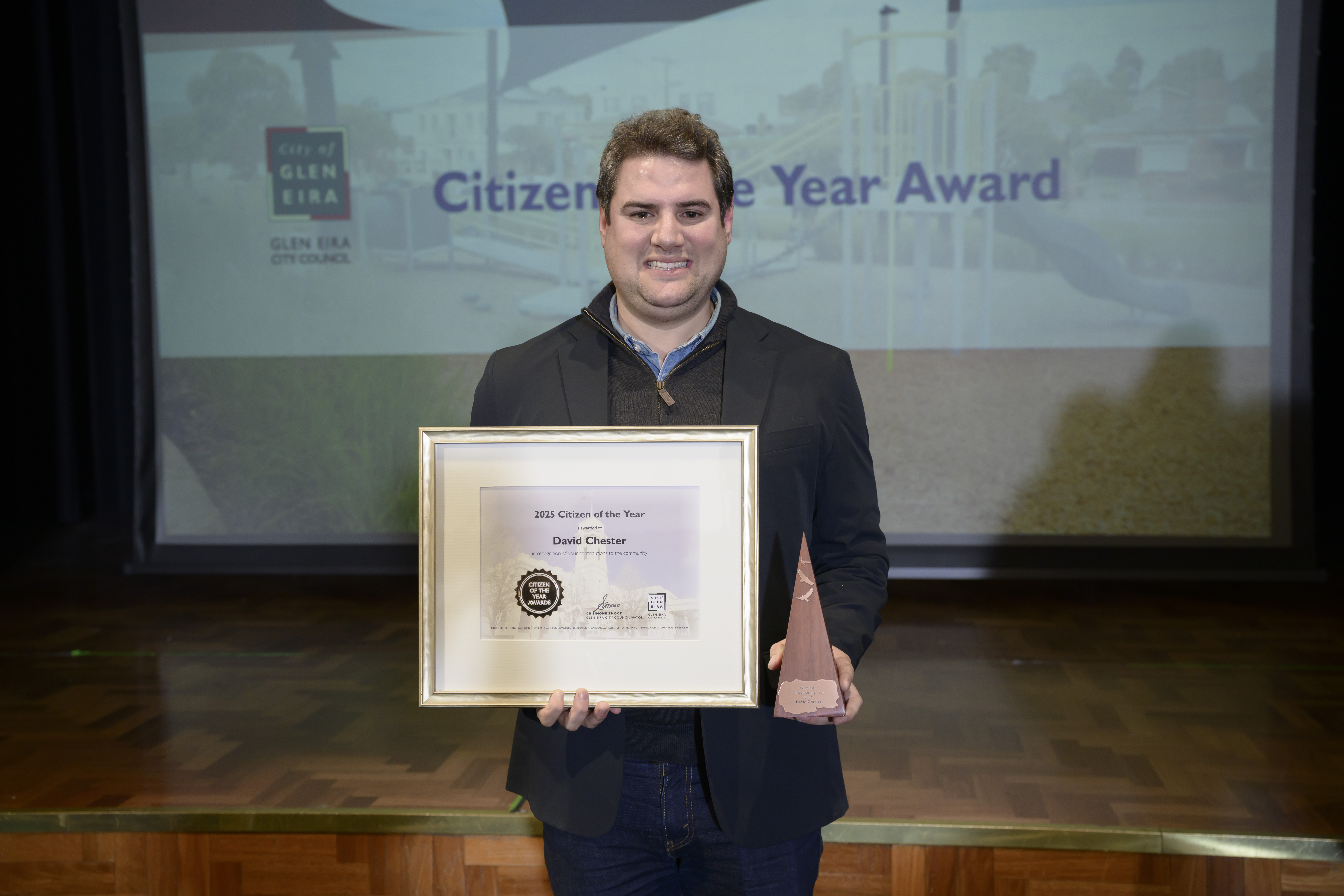 A person standing on a stage holding a framed certificate and a triangular award trophy. A large screen behind them displays the text “Citizen of the Year Award” with the City of Glen Eira branding.