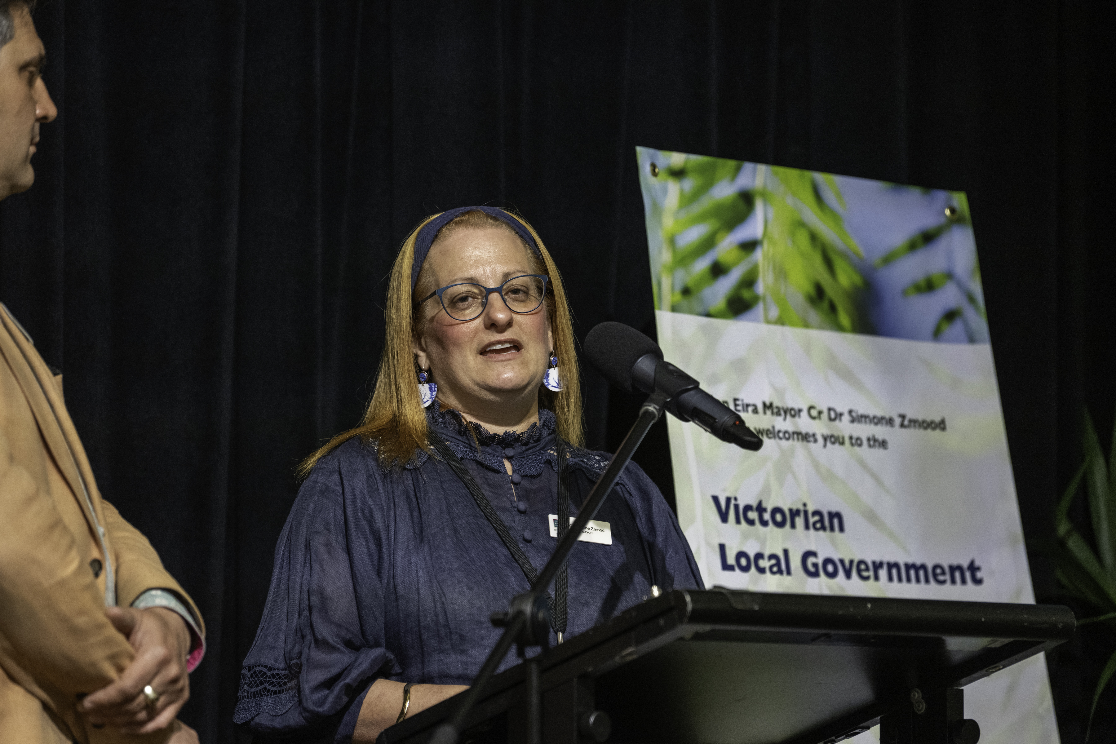 Person wearing glasses stands at a lectern speaking into a microphone at an event, with a sign behind them reading “Victorian Local Government” and another person partially visible to the side.