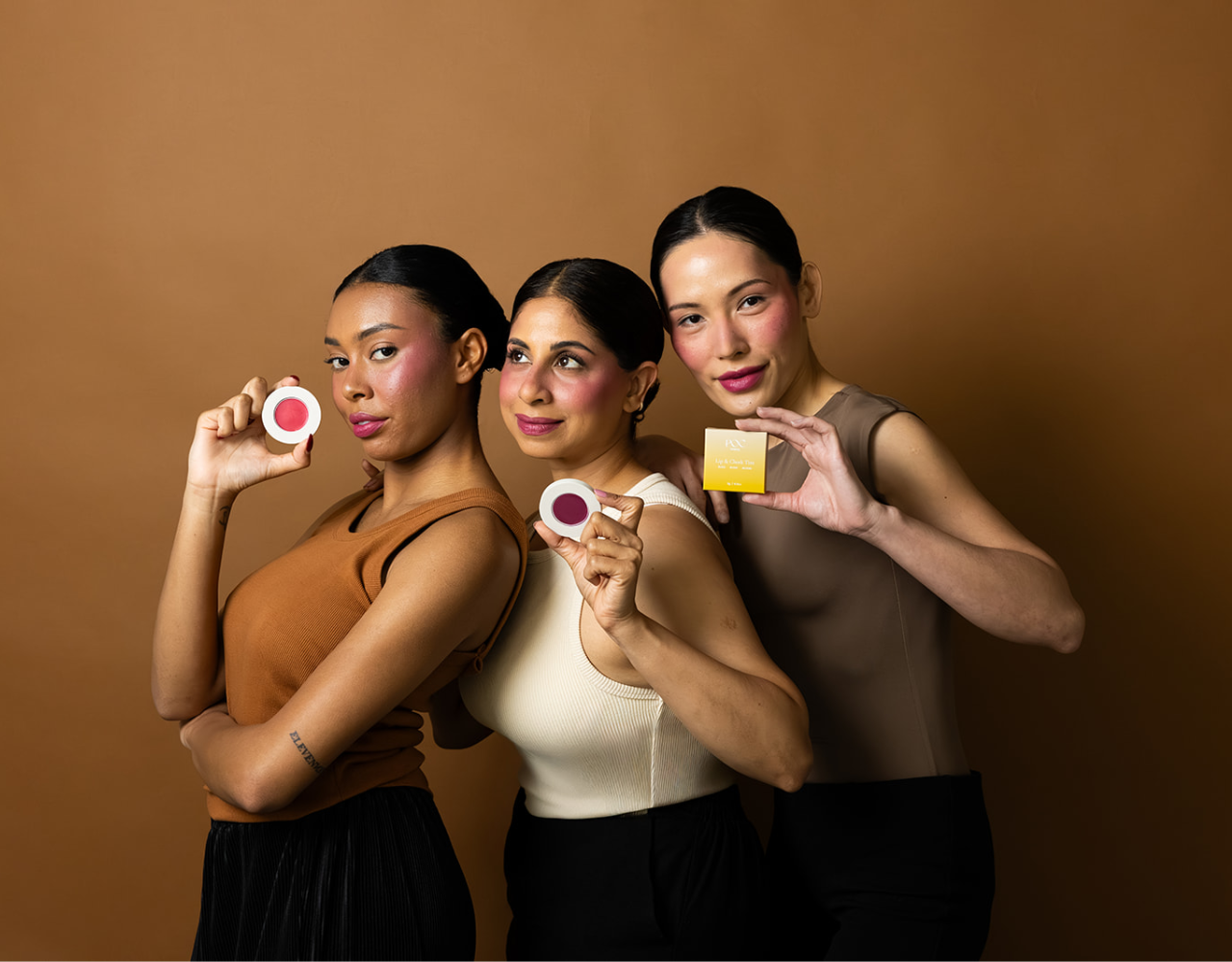 Three people pose against a warm brown background, each holding a POC Beauty cosmetic product. Two hold round makeup compacts and one holds a boxed product. The image showcases a range of beauty products and features a variety of people wearing neutral-toned clothing.