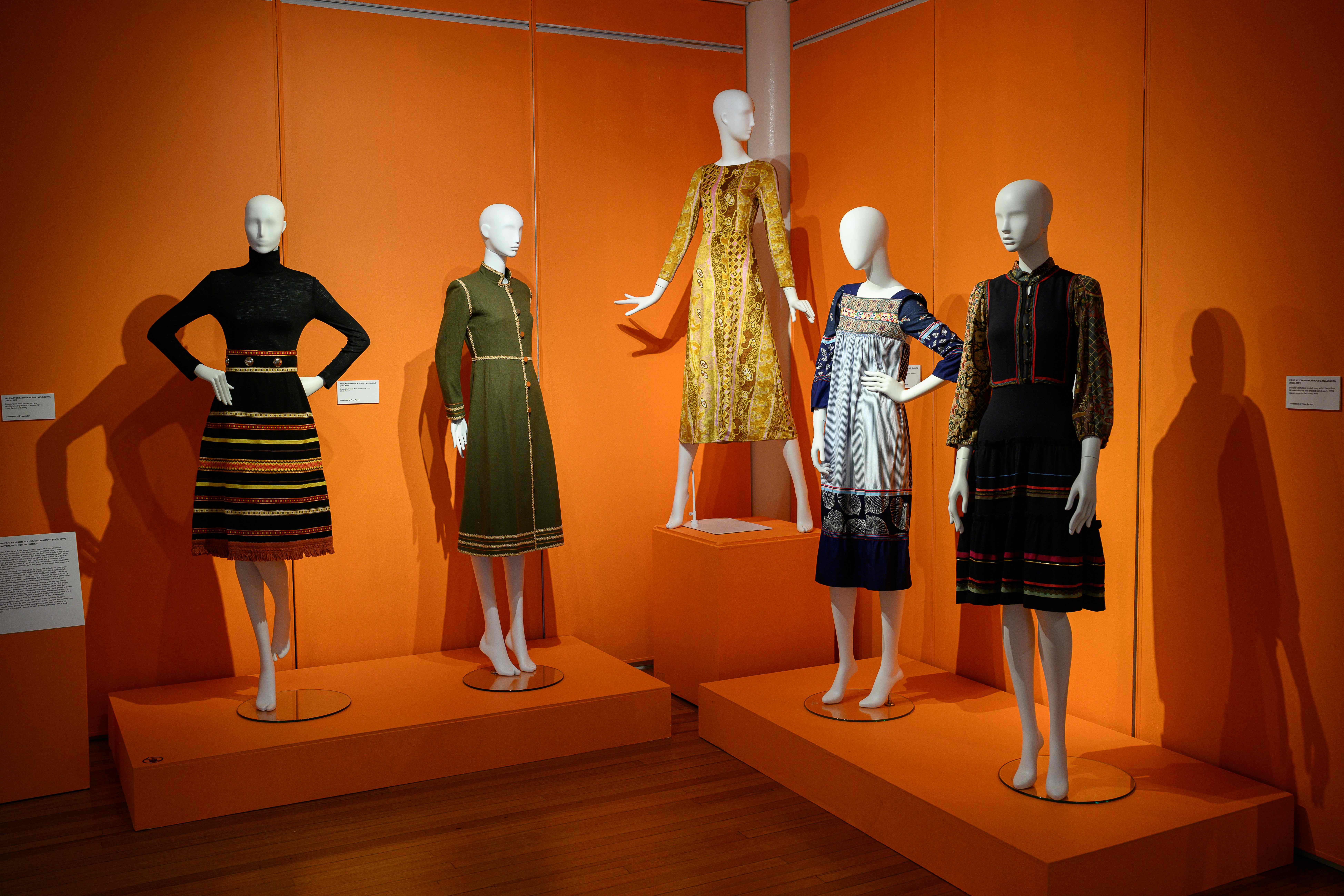Five mannequins display patterned dresses against bright orange walls in a fashion exhibition.