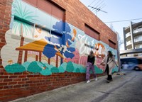 Two people walk past a colourful mural painted on a red brick wall in an urban laneway. The mural features stylised trees, birds, a pavilion, and large flowing lettering, with a bus passing by in the background.