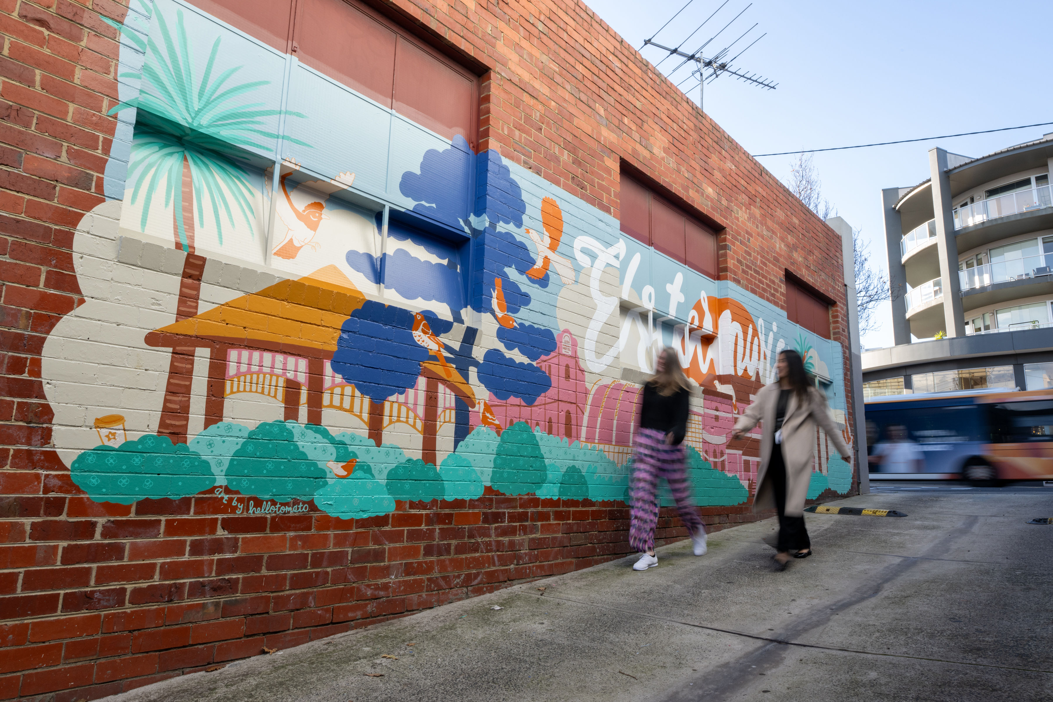 Two people walk past a colourful mural painted on a red brick wall in an urban laneway. The mural features stylised trees, birds, a pavilion, and large flowing lettering, with a bus passing by in the background.