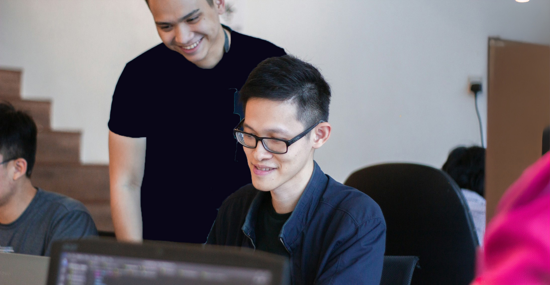 Two colleagues in an office setting smiling as they look at a computer screen together, one seated wearing glasses and the other standing beside him, suggesting collaboration and teamwork.