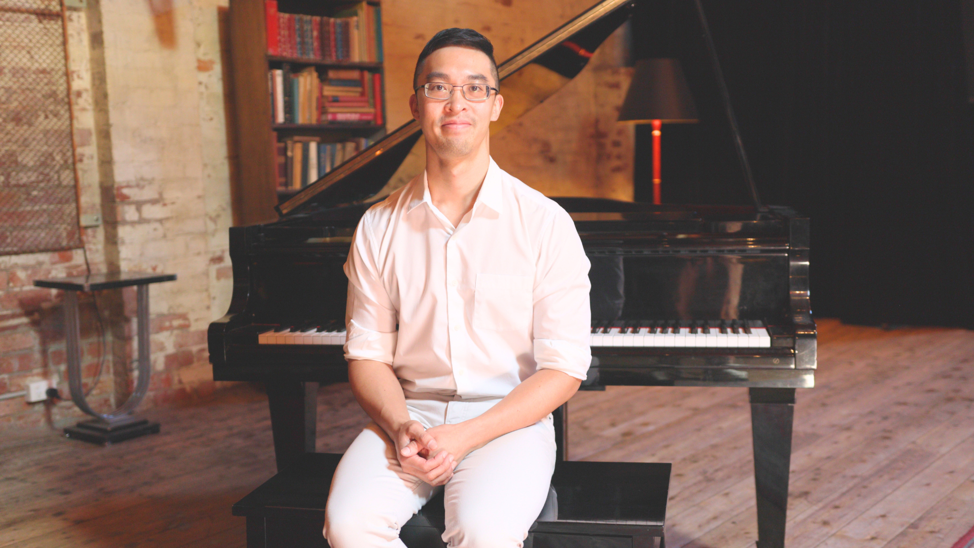 Person seated on a bench in front of a grand piano in a warmly lit studio, with bookshelves and a floor lamp in the background.