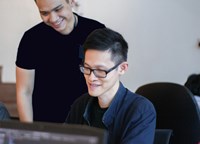 Two colleagues in an office setting smiling as they look at a computer screen together, one seated wearing glasses and the other standing beside him, suggesting collaboration and teamwork.
