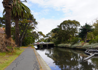 Pathway alongside Elster Creek showing calm water, surrounding trees and a stormwater litter trap near a small road bridge.