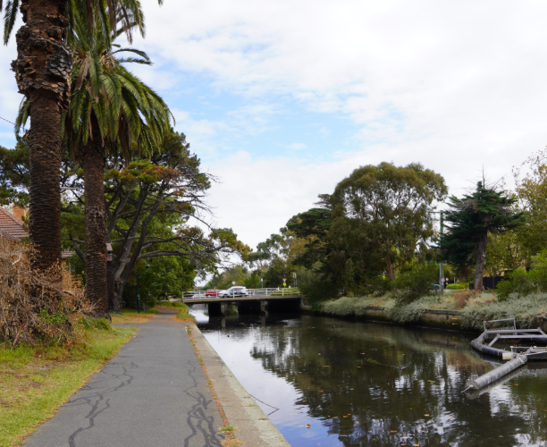 Pathway alongside Elster Creek showing calm water, surrounding trees and a stormwater litter trap near a small road bridge.