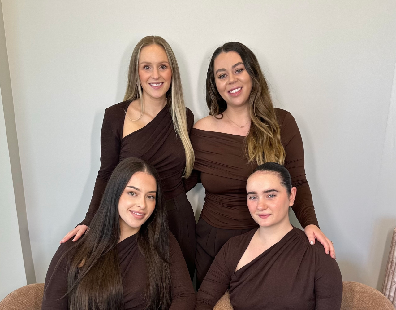 Four team members pose together in a salon or studio setting. Two people are seated in armchairs while two stand behind them, with each person resting a hand on a colleague’s shoulder. All wear coordinated dark brown tops against a light grey background.