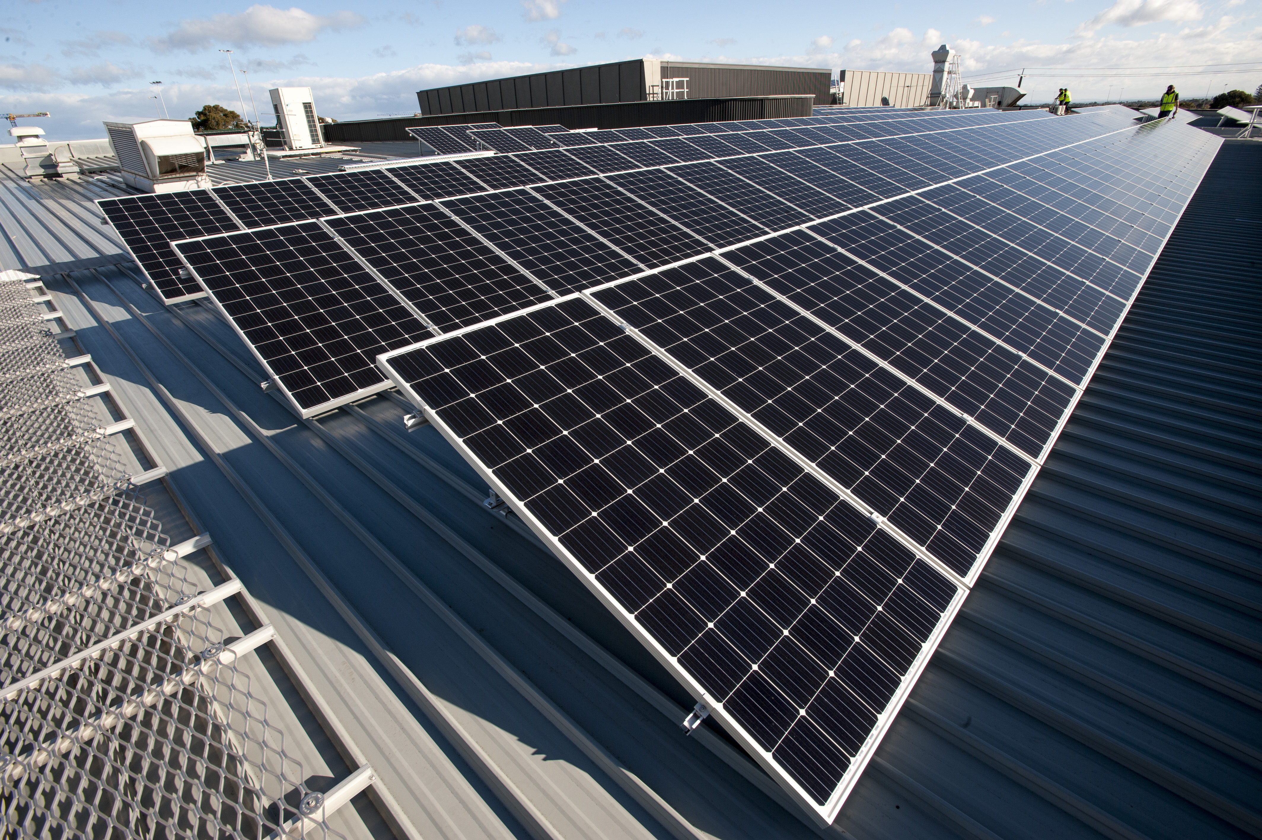 Rows of solar panels installed across a large metal rooftop, angled toward the sun, with industrial buildings and two workers visible in the distance under a partly cloudy sky.