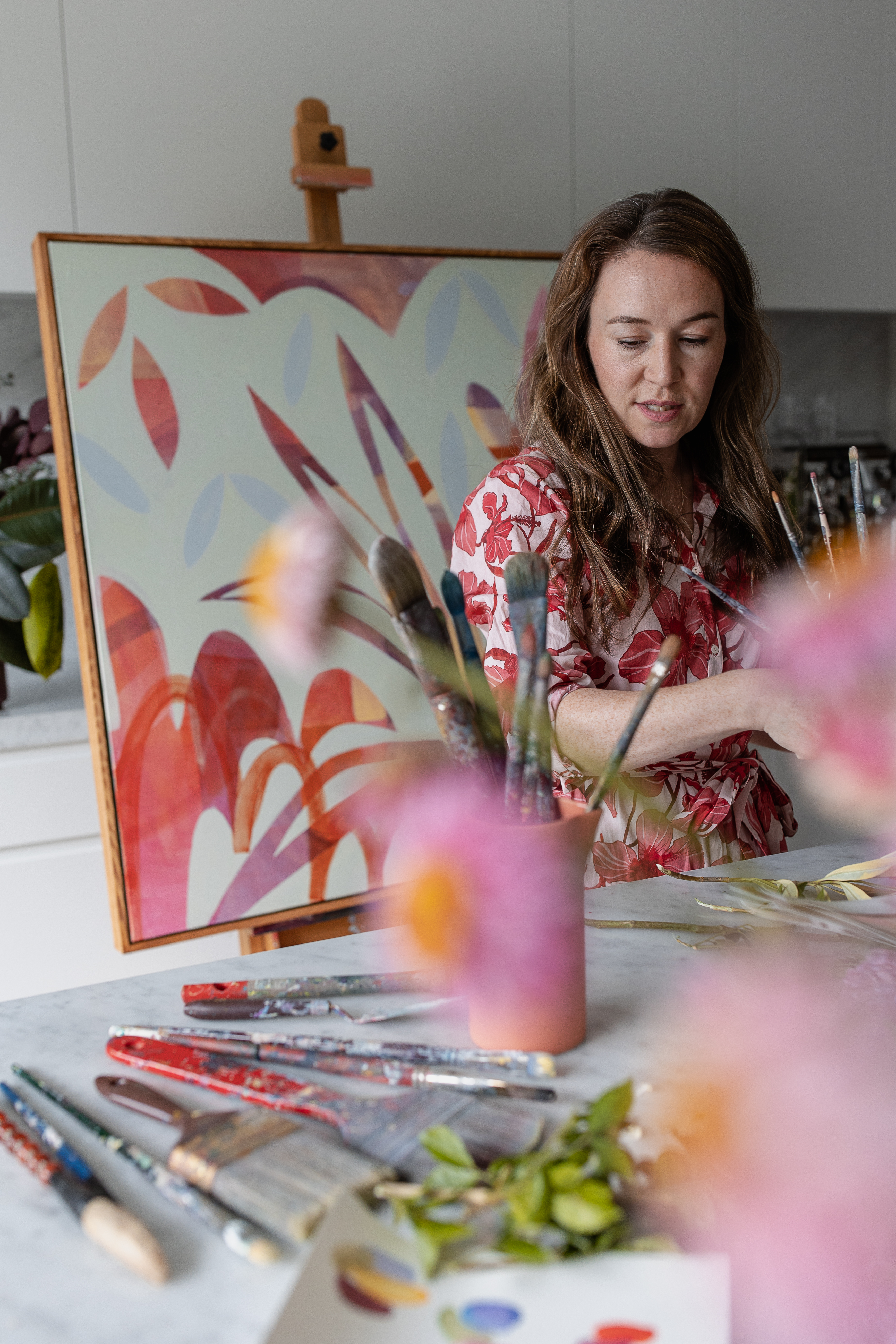 Person working at a table with paintbrushes and materials, with an abstract painting on an easel behind them.