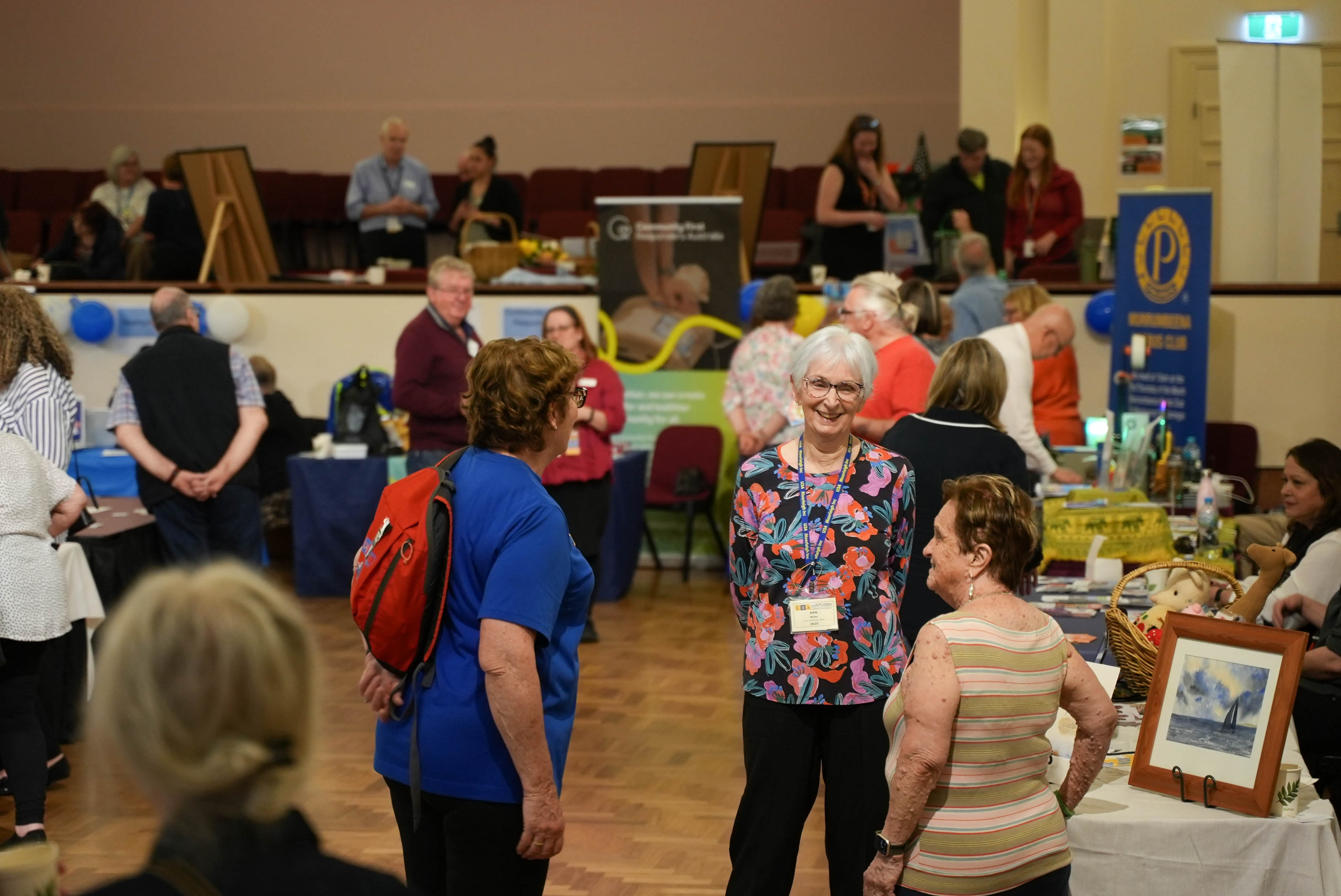 People browse information stalls and chat with exhibitors at a Healthy Ageing Expo in a community hall.