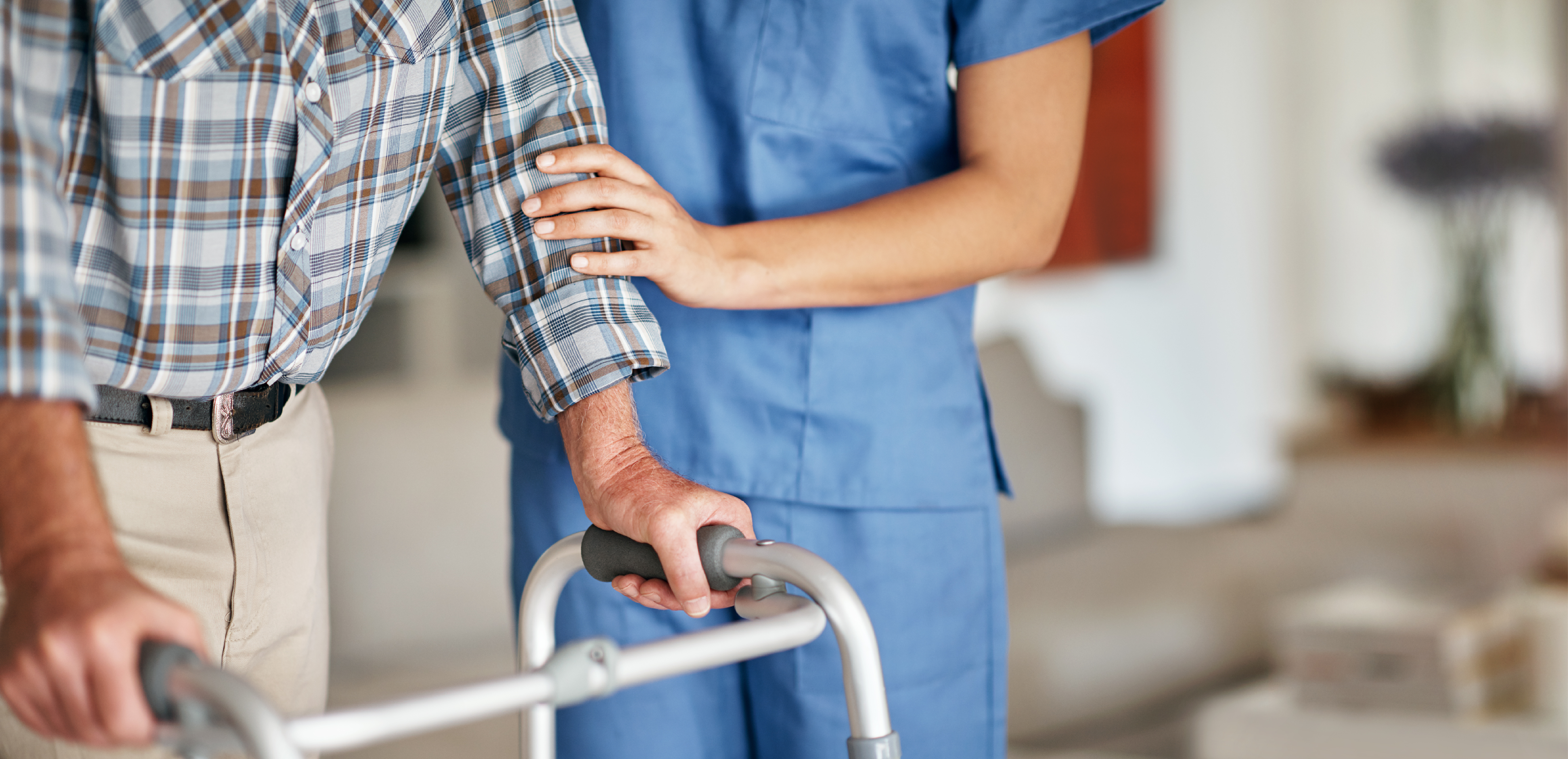 Close-up of an older person using a walking frame while a healthcare worker in blue scrubs stands beside them with a supportive hand on their arm in a home setting.