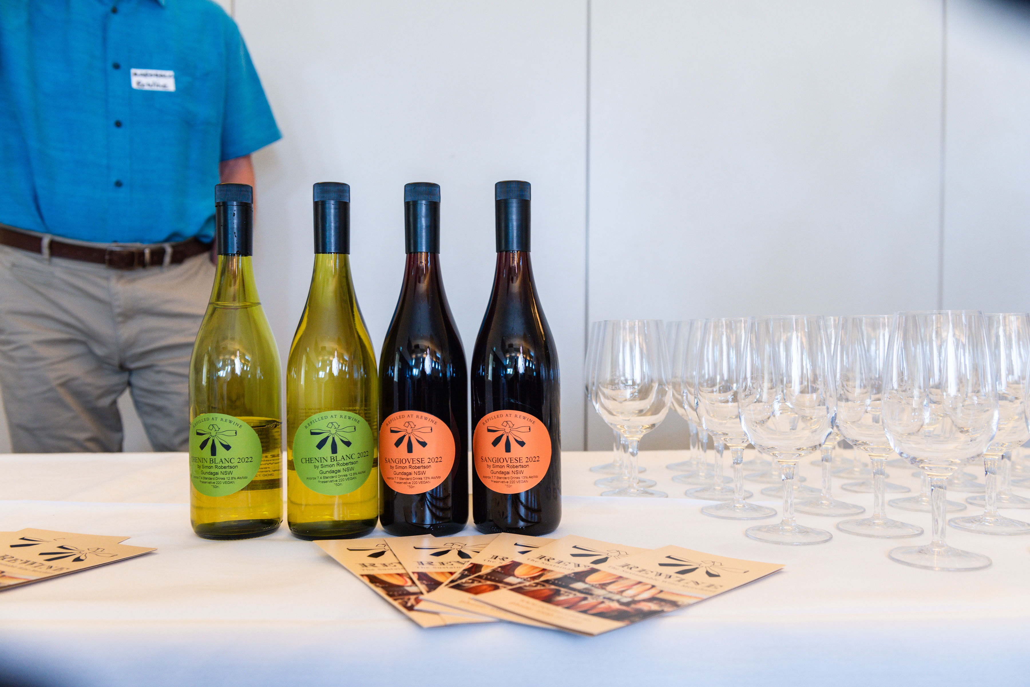 Four wine bottles and rows of empty tasting glasses displayed on a table with promotional cards.