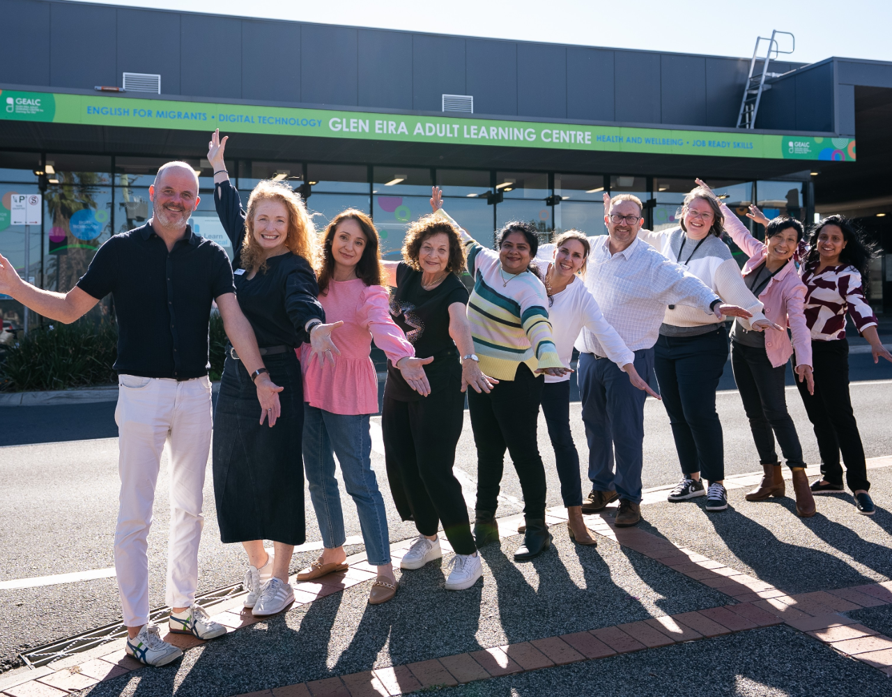A diverse group of staff members stand outside the Glen Eira Adult Learning Centre with arms outstretched in a welcoming pose. The group is gathered in front of the centre's entrance beneath signage promoting English for Migrants, Digital Technology, Health and Wellbeing, and Job Ready Skills programs.