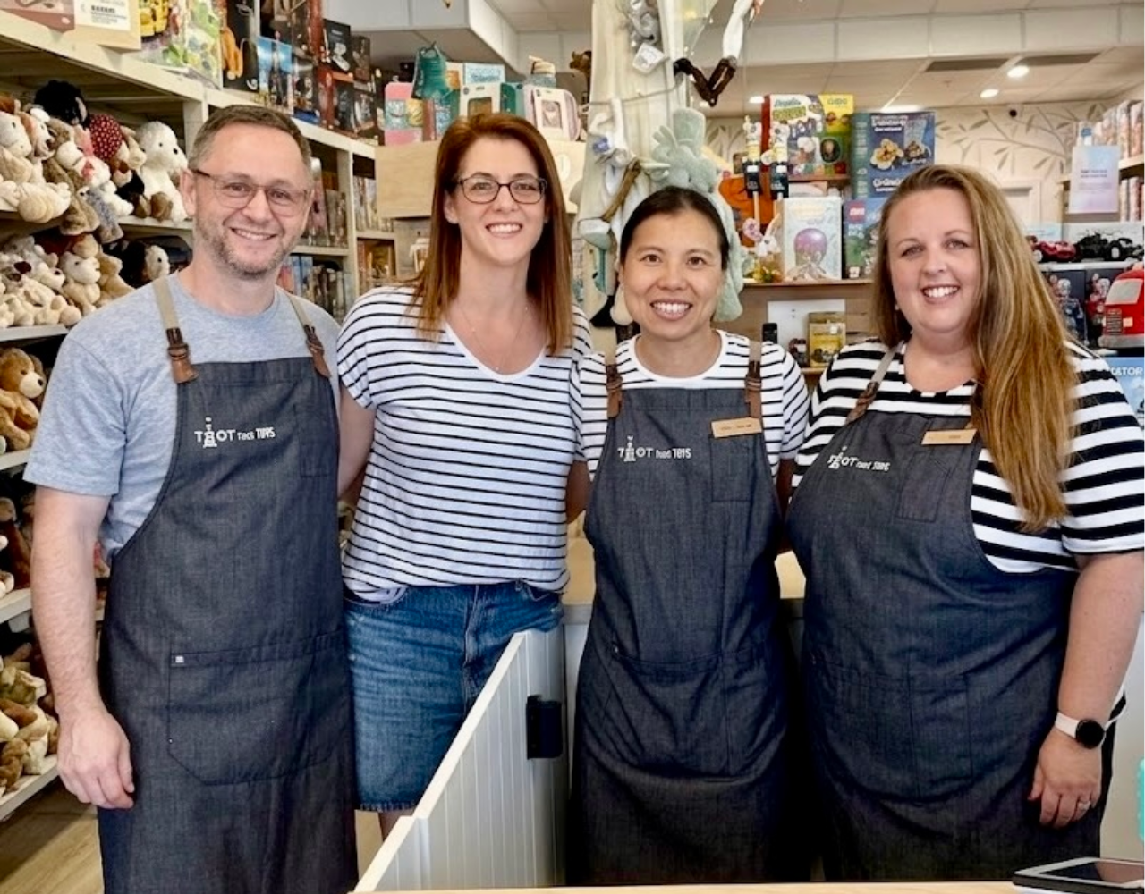 Four staff members stand together behind a counter inside a toy store. Some wear striped shirts and branded aprons, with shelves of toys, games and stuffed animals visible in the background.