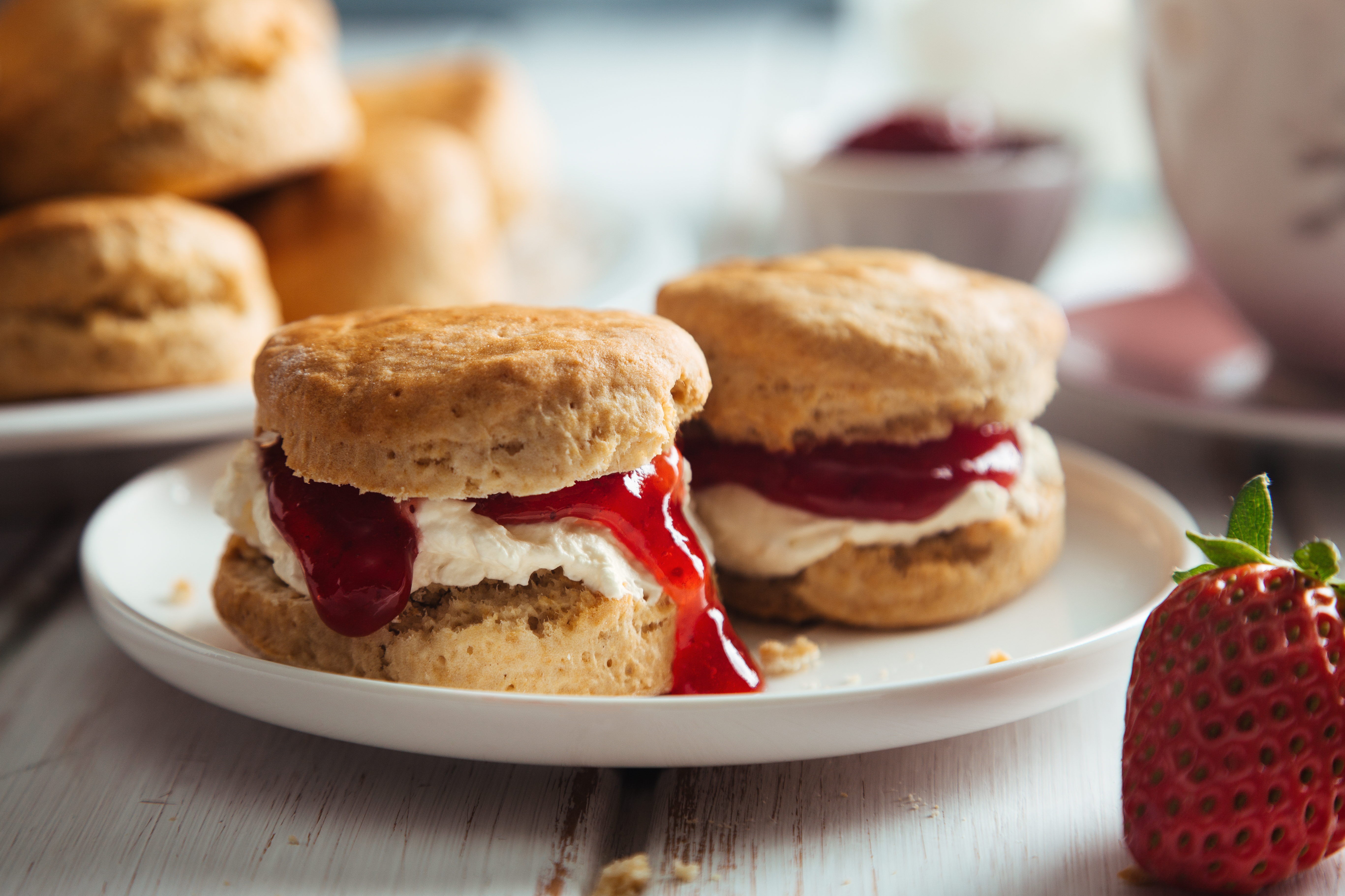 Freshly baked scones filled with cream and strawberry jam on a plate, with strawberries and extra scones in the background.