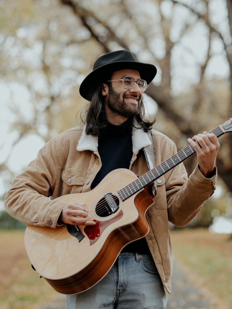A man in an autumnal setting playing guitar. He has a black hat on, winter jacket and is wearing glasses.