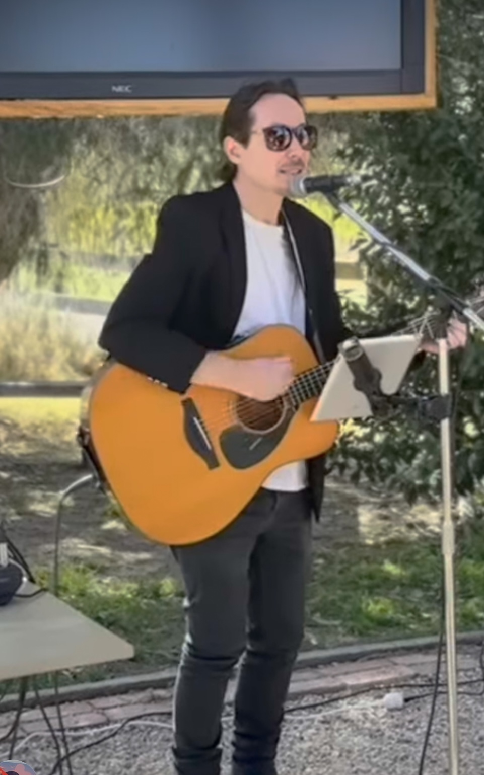 Man playing an acoustic guitar and singing into a microphone during an outdoor live music performance.