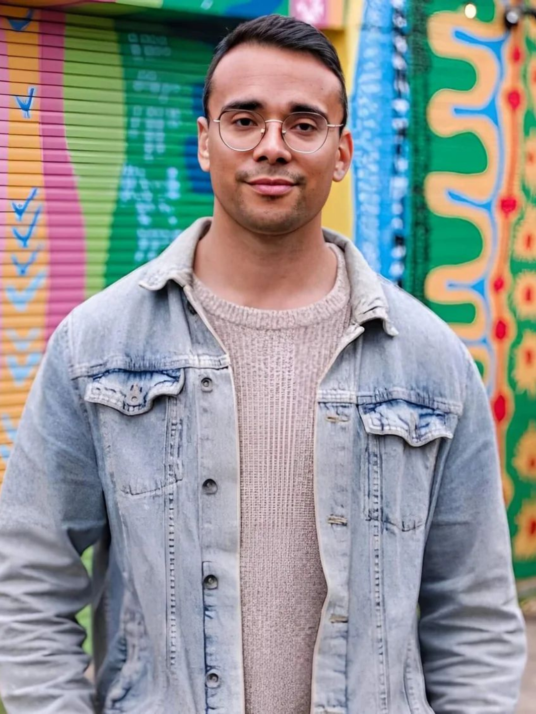 A man with glasses and a denim jacket is looking at camera. Background is a colourful wall.