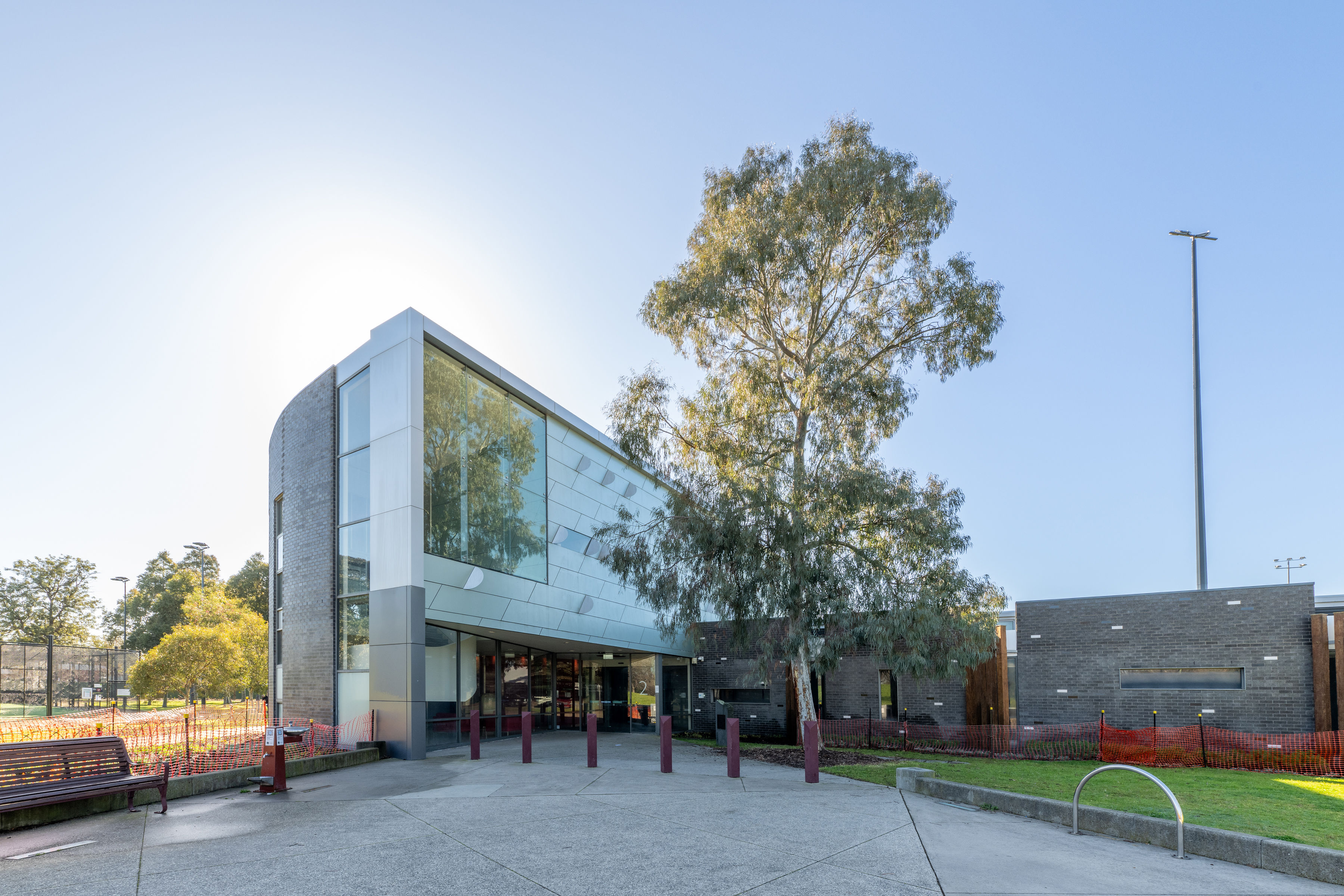 A modern building with large glass windows and a tall tree in front, surrounded by paved walkways, benches, and landscaped areas on a sunny day.