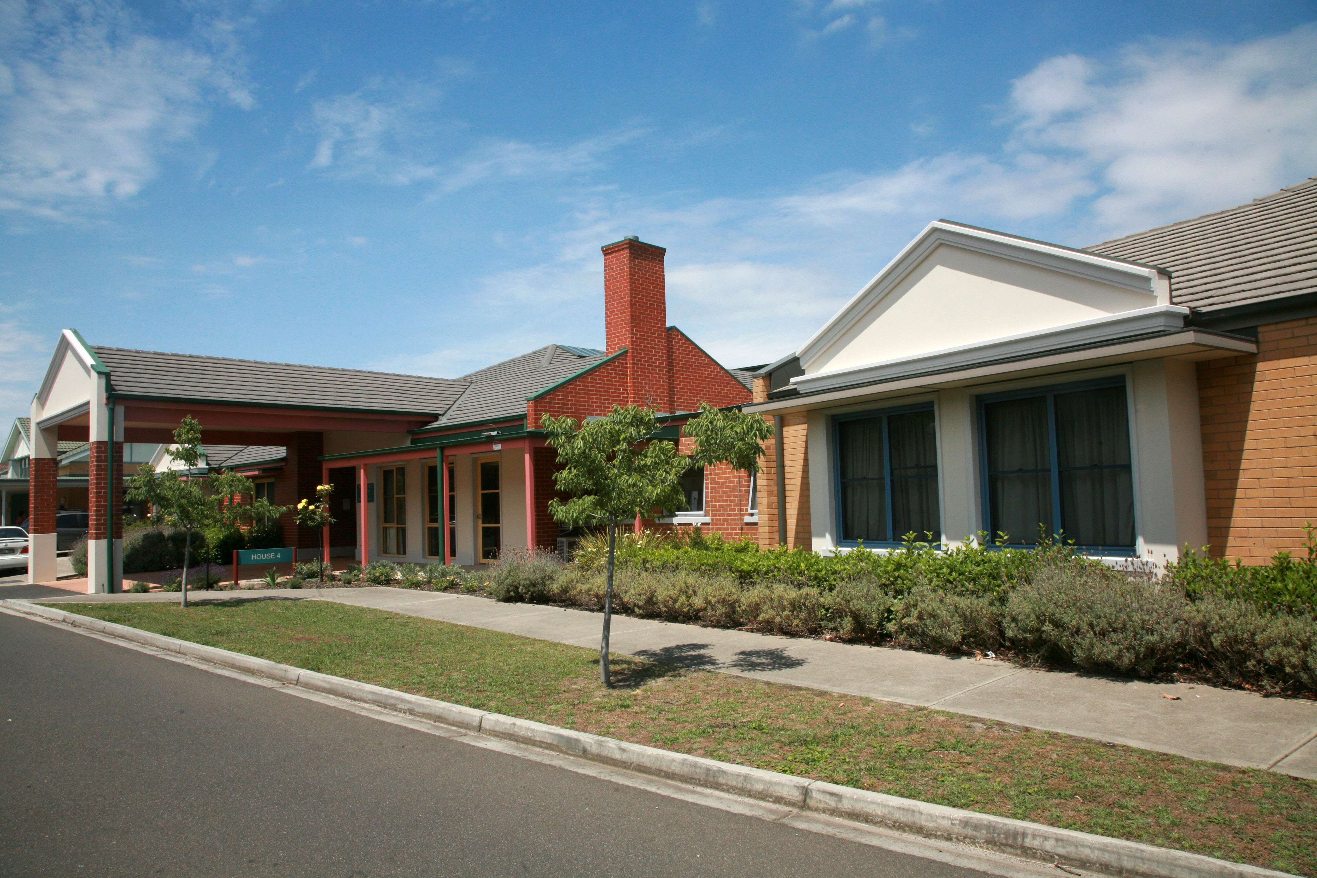 Modern single-storey residential care facility with red brick and light-rendered buildings, a covered entrance, landscaped garden beds and small trees along a suburban street under a blue sky.