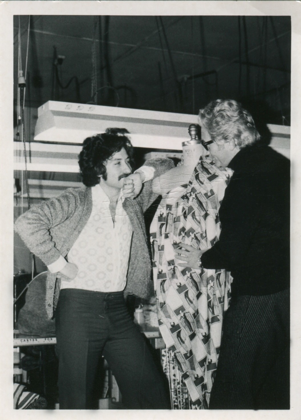 Black-and-white archival photo showing two people in a clothing workshop adjusting a dress on a mannequin, with sewing equipment and work surfaces visible in the background.