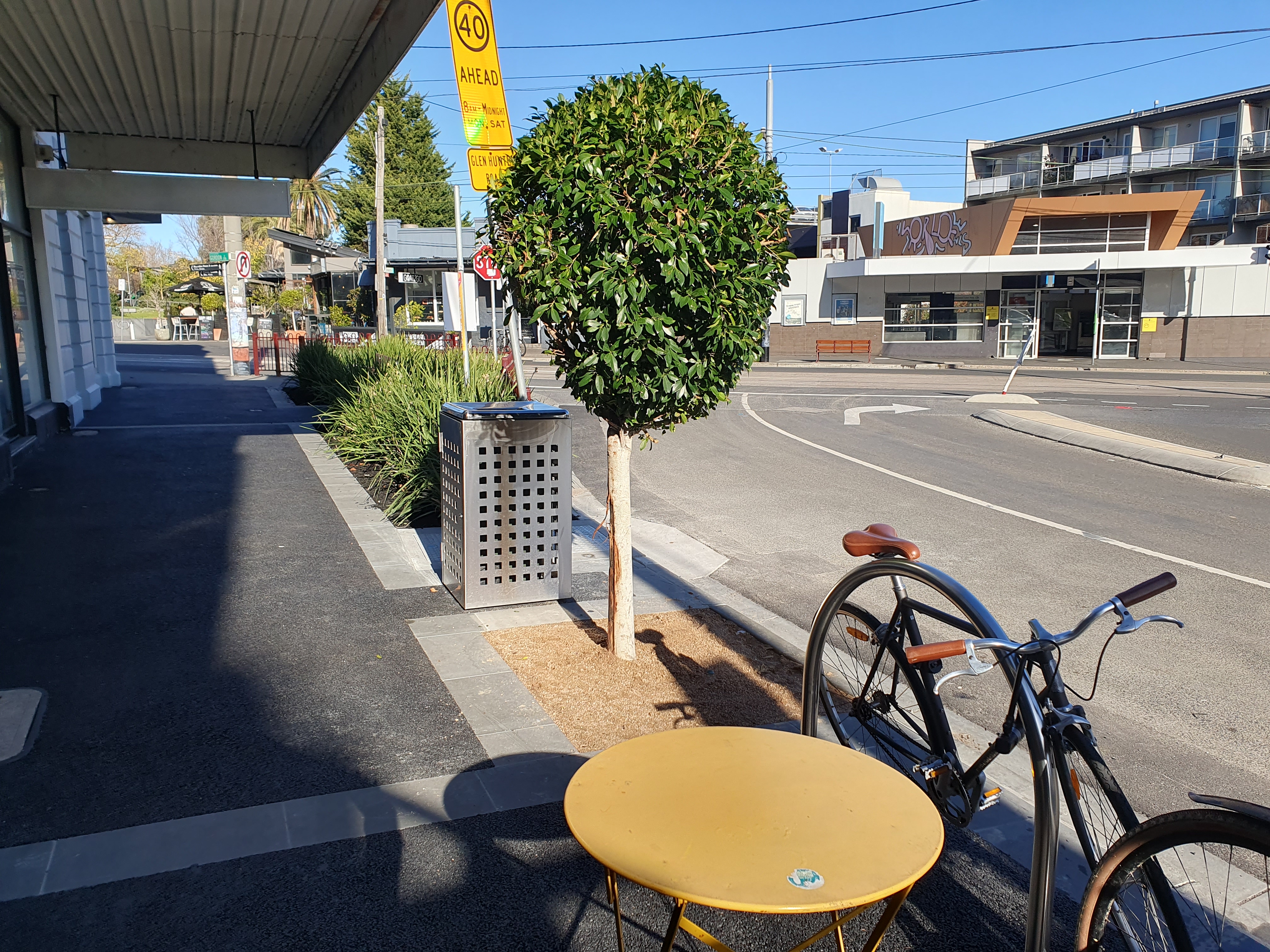 Streetscape with a bicycle parked beside a small outdoor table on a wide footpath, facing a quiet shopping strip and road intersection.
