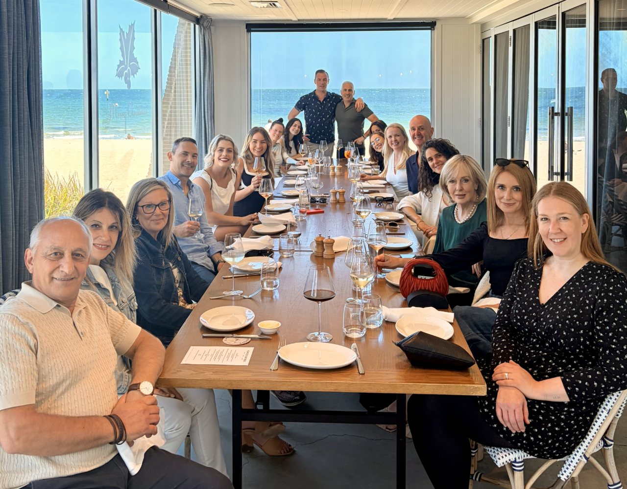 A large group of people sit around a long dining table at a beachfront restaurant. Plates, glasses and wine glasses are arranged on the table, with floor-to-ceiling windows revealing a sandy beach and ocean view in the background. Two people stand at the far end of the table while the group faces the camera.