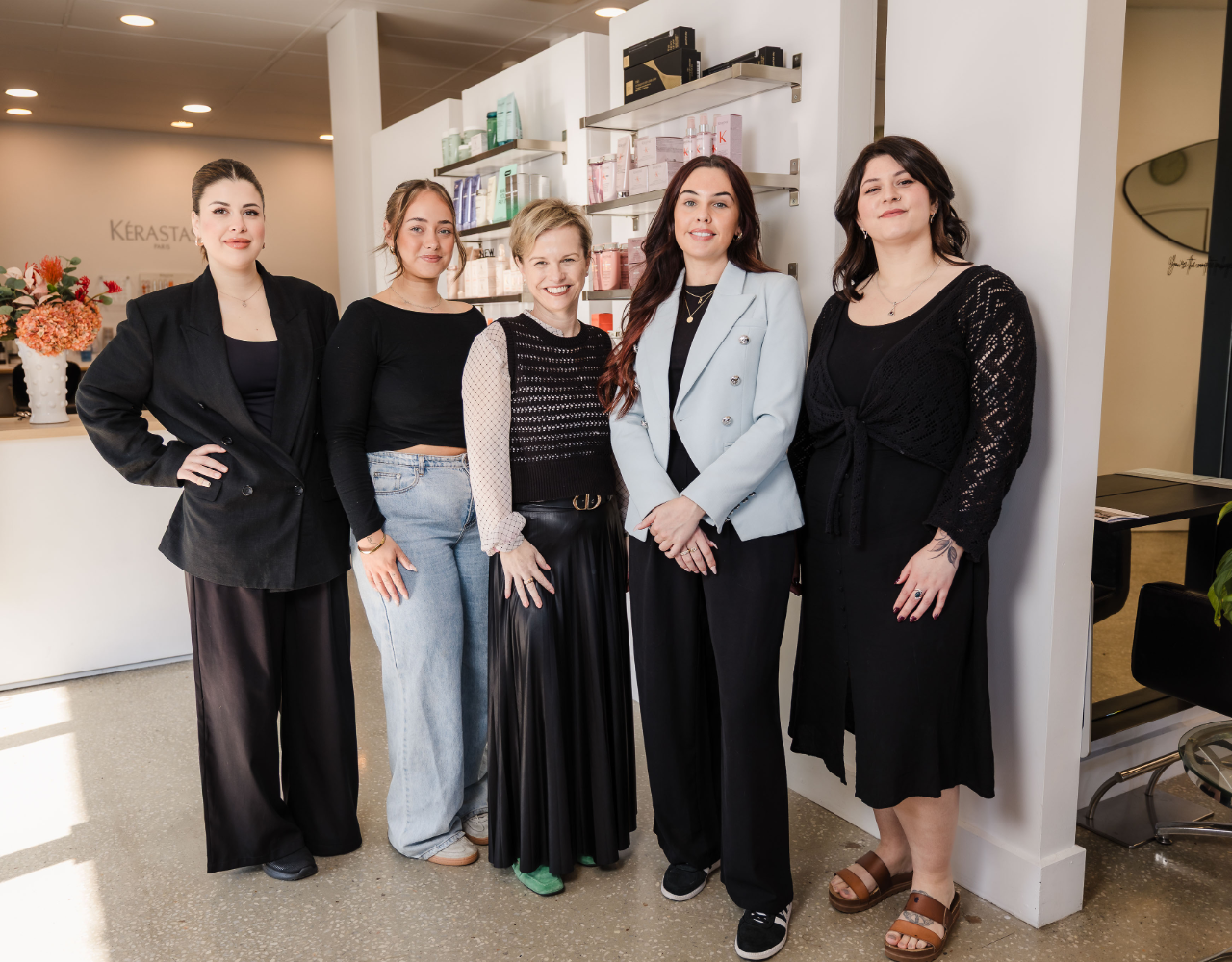 Five team members stand together inside a salon reception area. Product shelves, salon stations and decorative flowers are visible in the bright, modern space behind them.