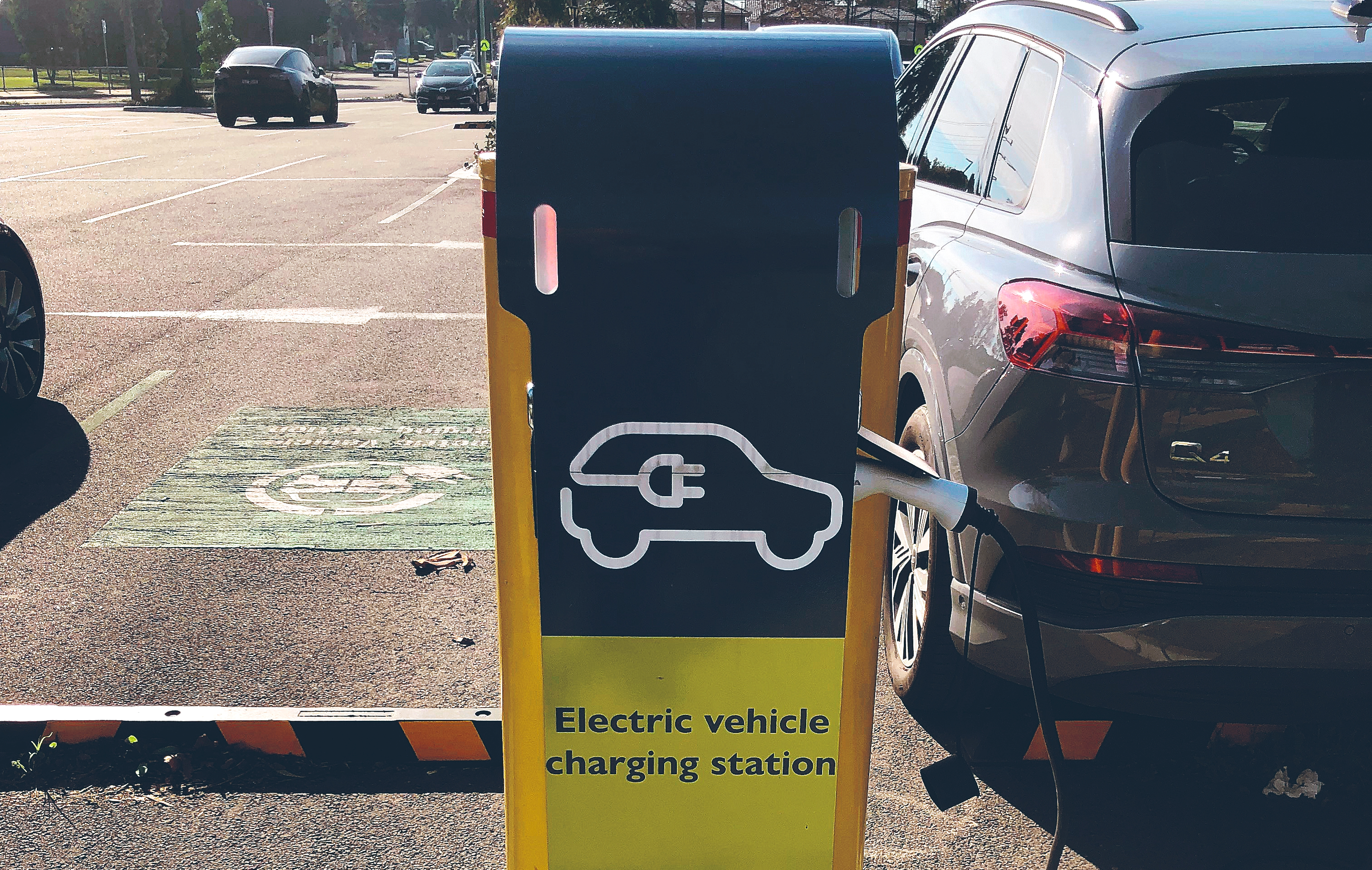 Public electric vehicle charging station beside a parked SUV, with a designated EV charging bay visible in the background.