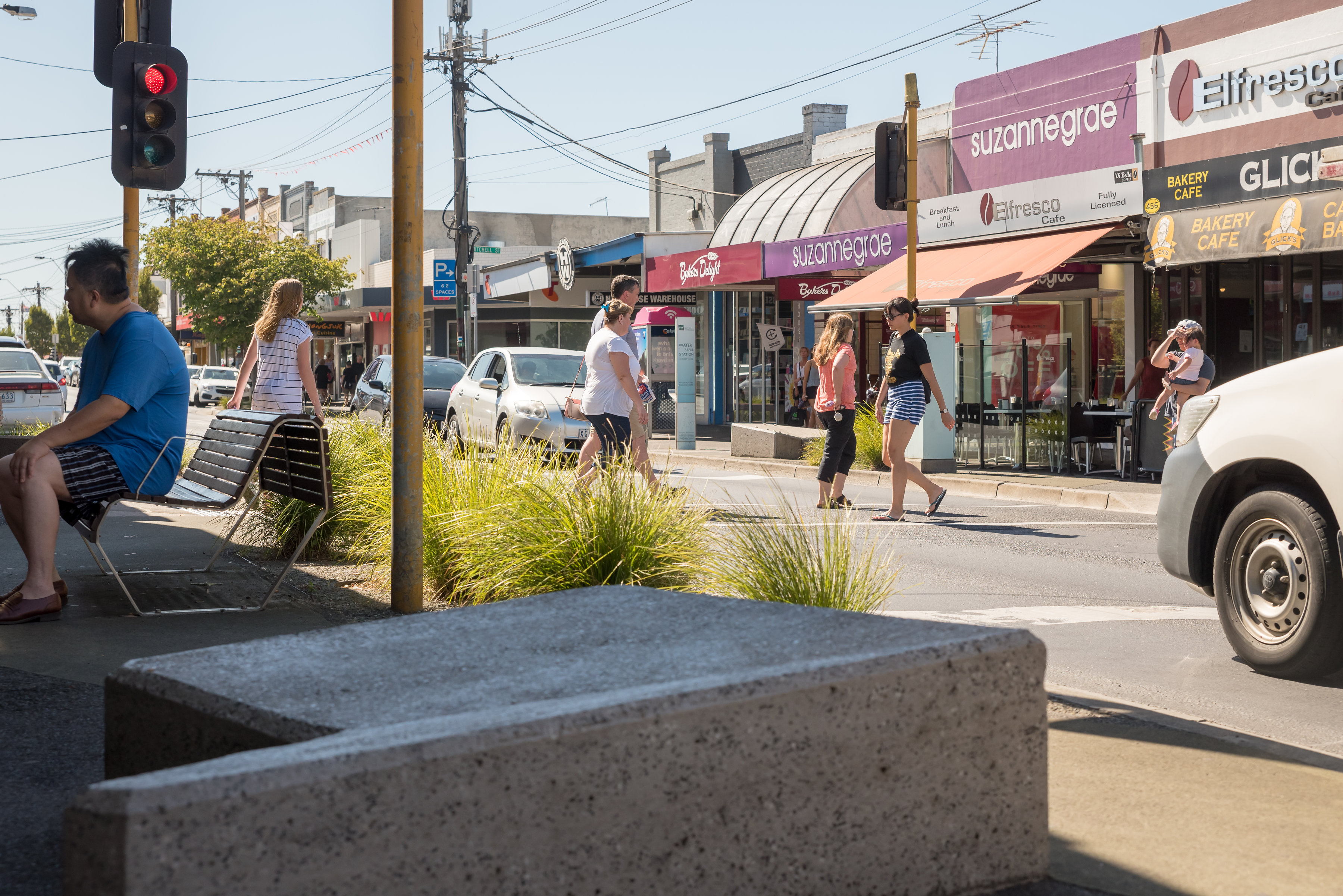 Street scene in Bentleigh showing pedestrians crossing at a signalised intersection, with shops and cafés along Centre Road and cars stopped at the traffic lights.