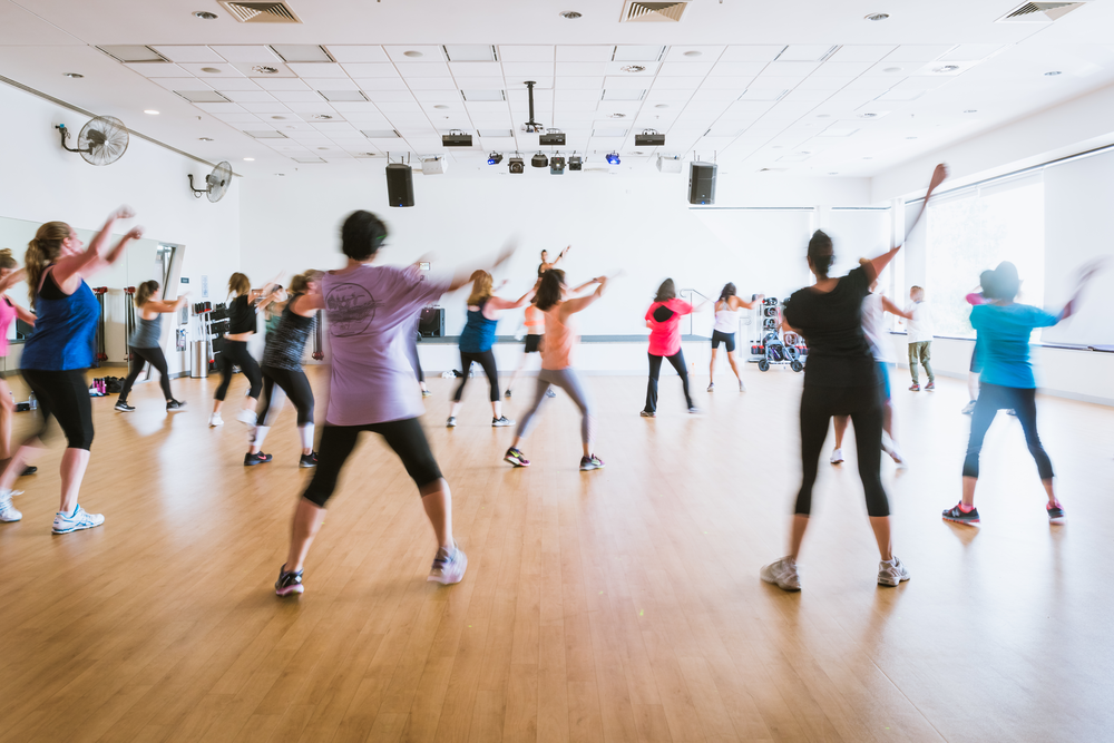 Participants take part in a Zumba Gold fitness class in a community hall, following an instructor-led dance workout.