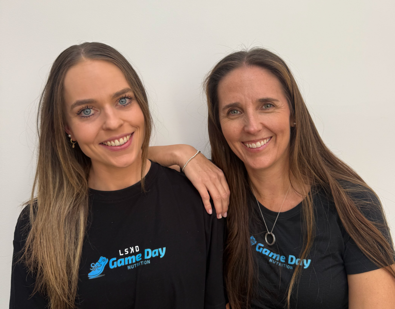 Two staff members from Game Day Nutrition stand side by side against a plain light-coloured background. Both wear black branded T-shirts featuring the Game Day Nutrition logo. One person rests an arm on the other's shoulder as they face the camera.