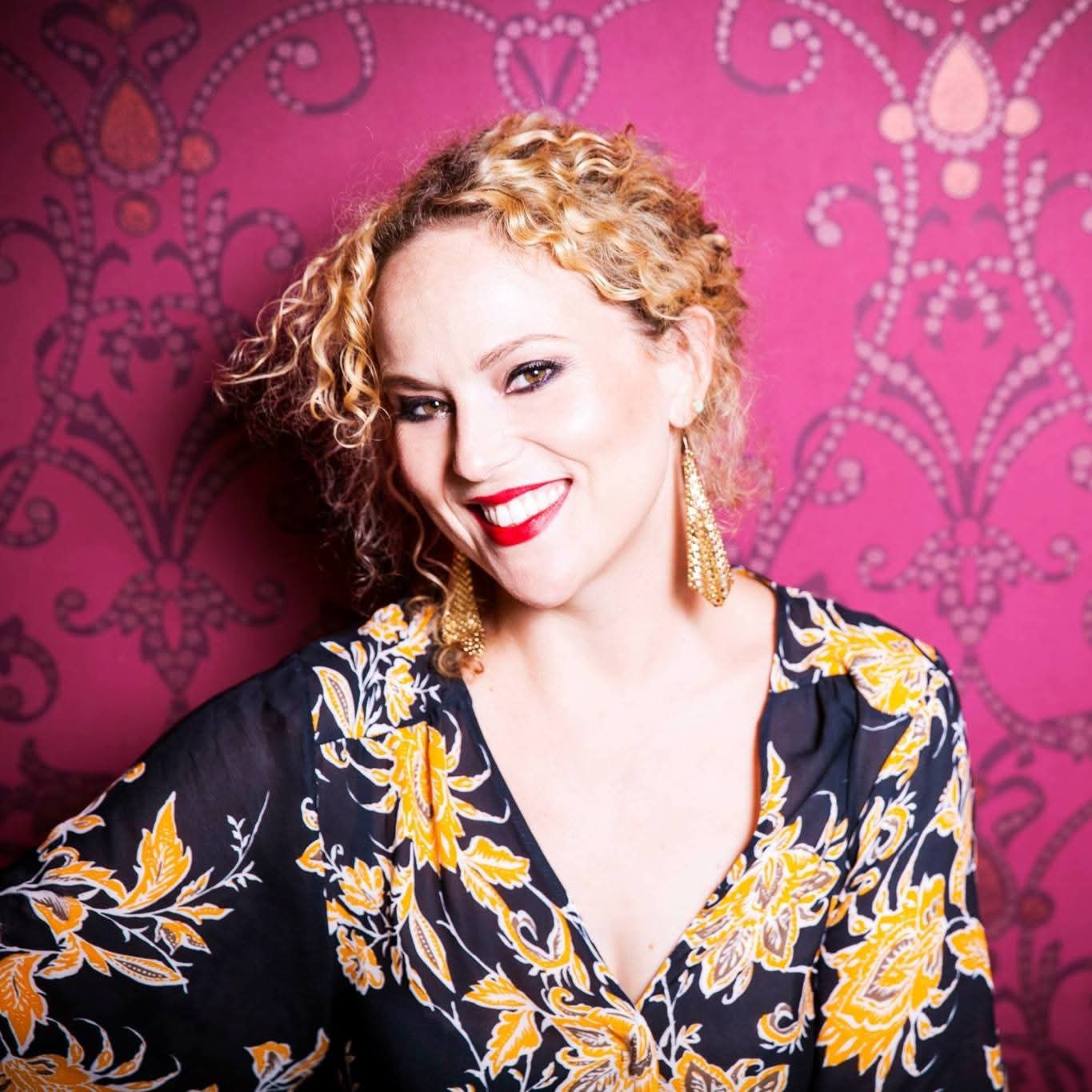 Portrait of a person with curly hair wearing a patterned top and large earrings, standing against a pink patterned background.
