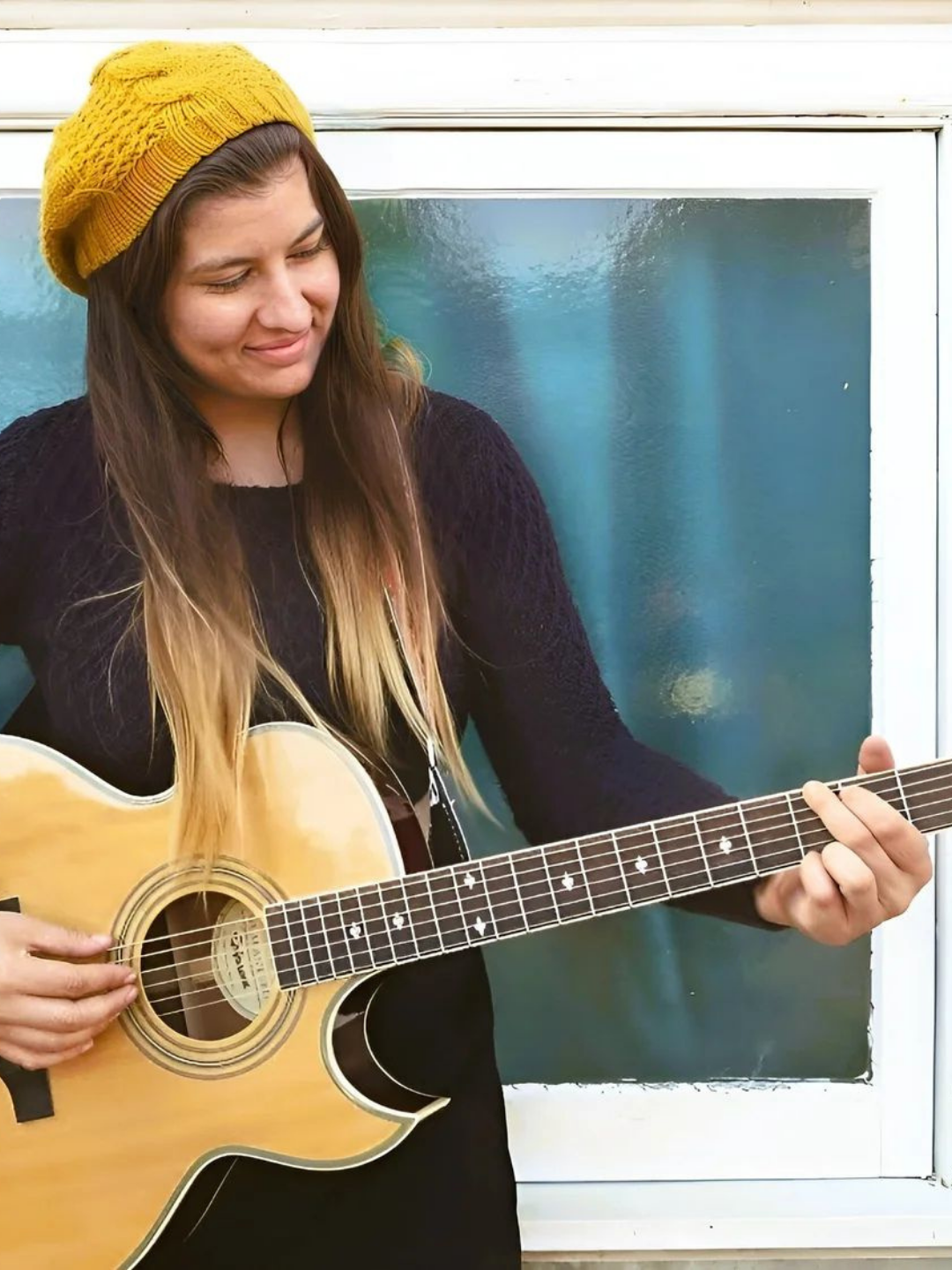 A lady with long brown hair and a yellow beanie, standing in front of a mirror and playing guitar. She is not looking at the camera.