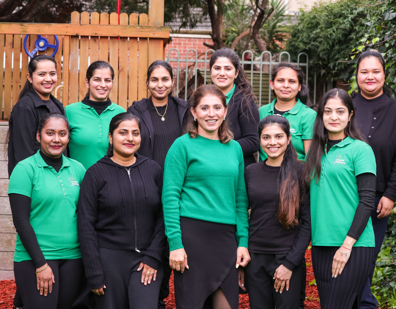 A group of childcare staff stand together outdoors in front of a playground and garden area. Several staff wear green branded polo shirts while others wear black clothing. Trees, climbing equipment and garden beds are visible in the background.