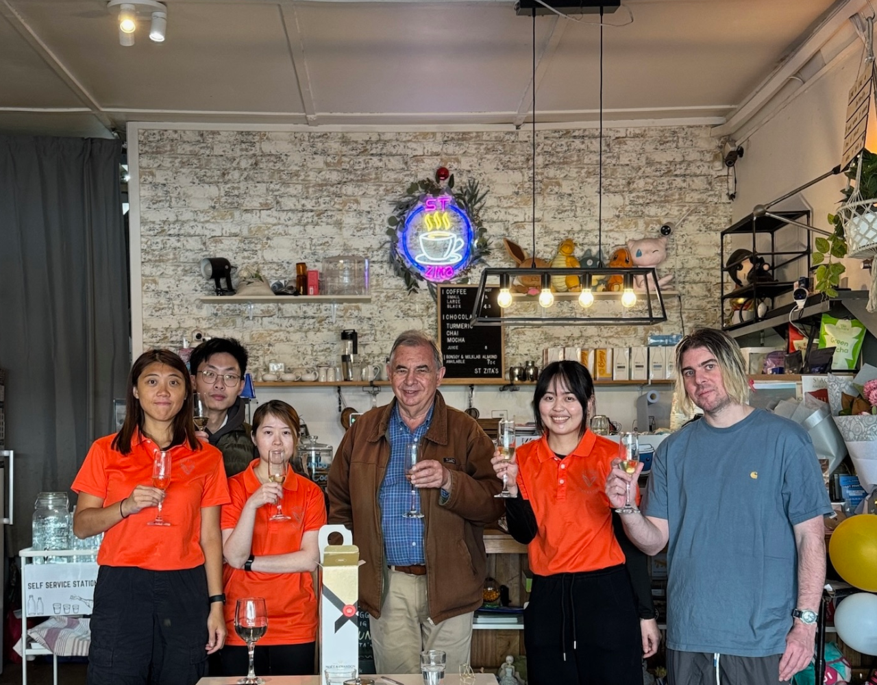 Six people stand inside a café holding champagne flutes for a toast. Three people wear bright orange shirts, while others wear casual clothing. The group stands in front of a white brick wall with shelves, café equipment and a neon coffee cup sign. A table with glasses and a gift box is in the foreground.