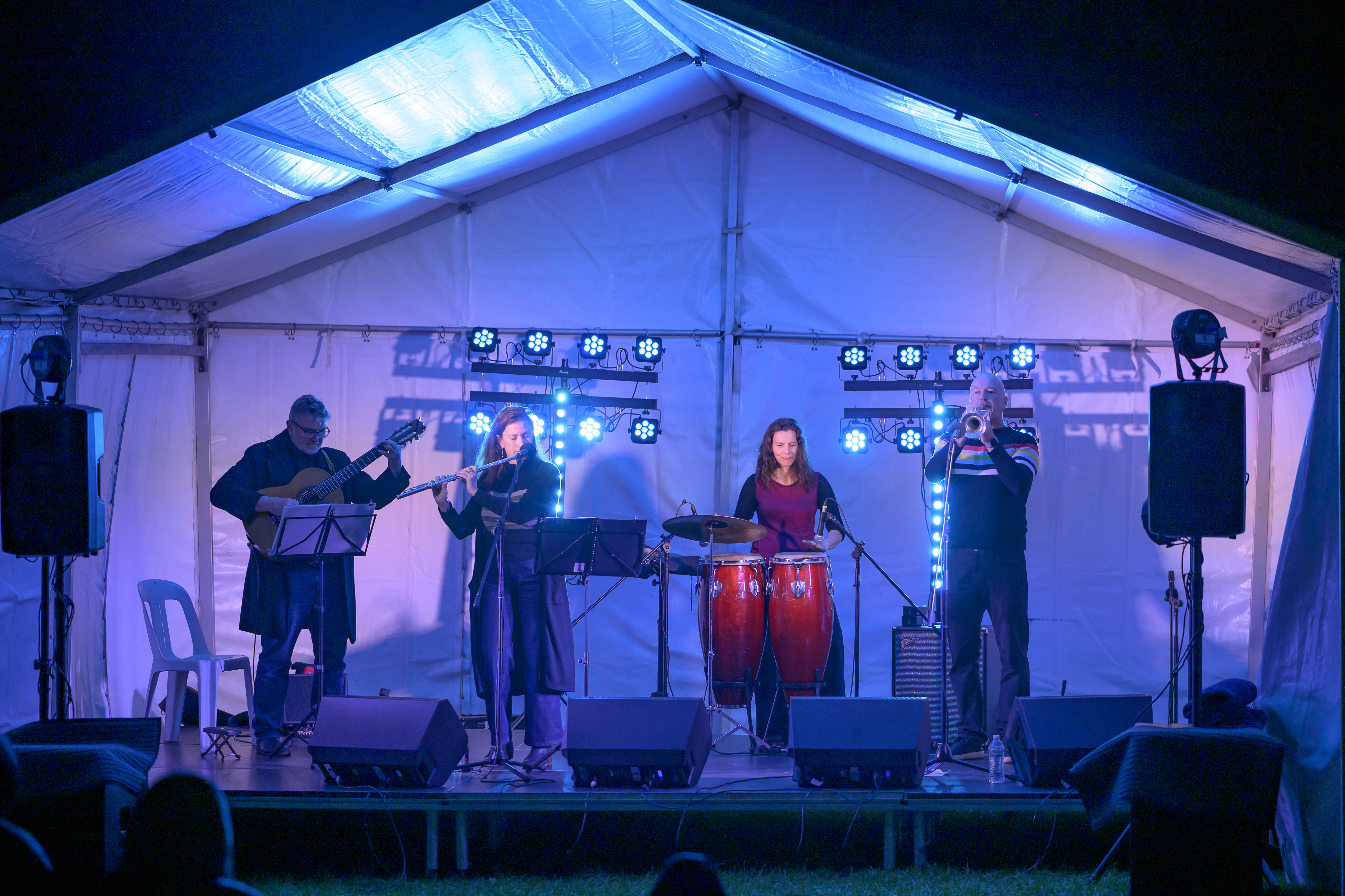 Small musical group performing on an outdoor stage under a white marquee at night, featuring guitar, flute, trumpet and percussion, with blue stage lighting, speakers and music stands visible.