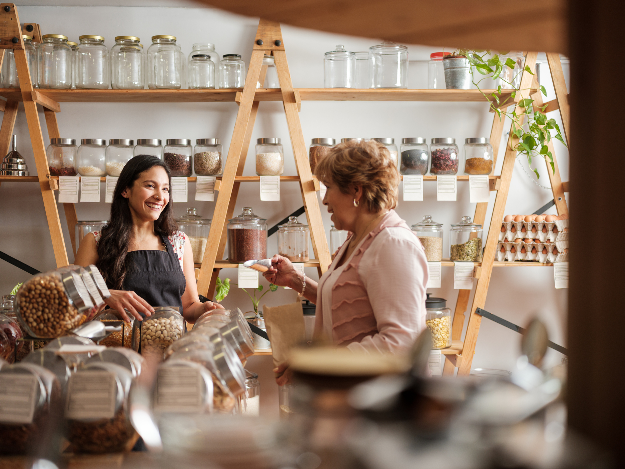 A shop assistant smiles while serving a customer at a bulk food store, surrounded by glass jars filled with grains, nuts and spices on wooden shelves.
