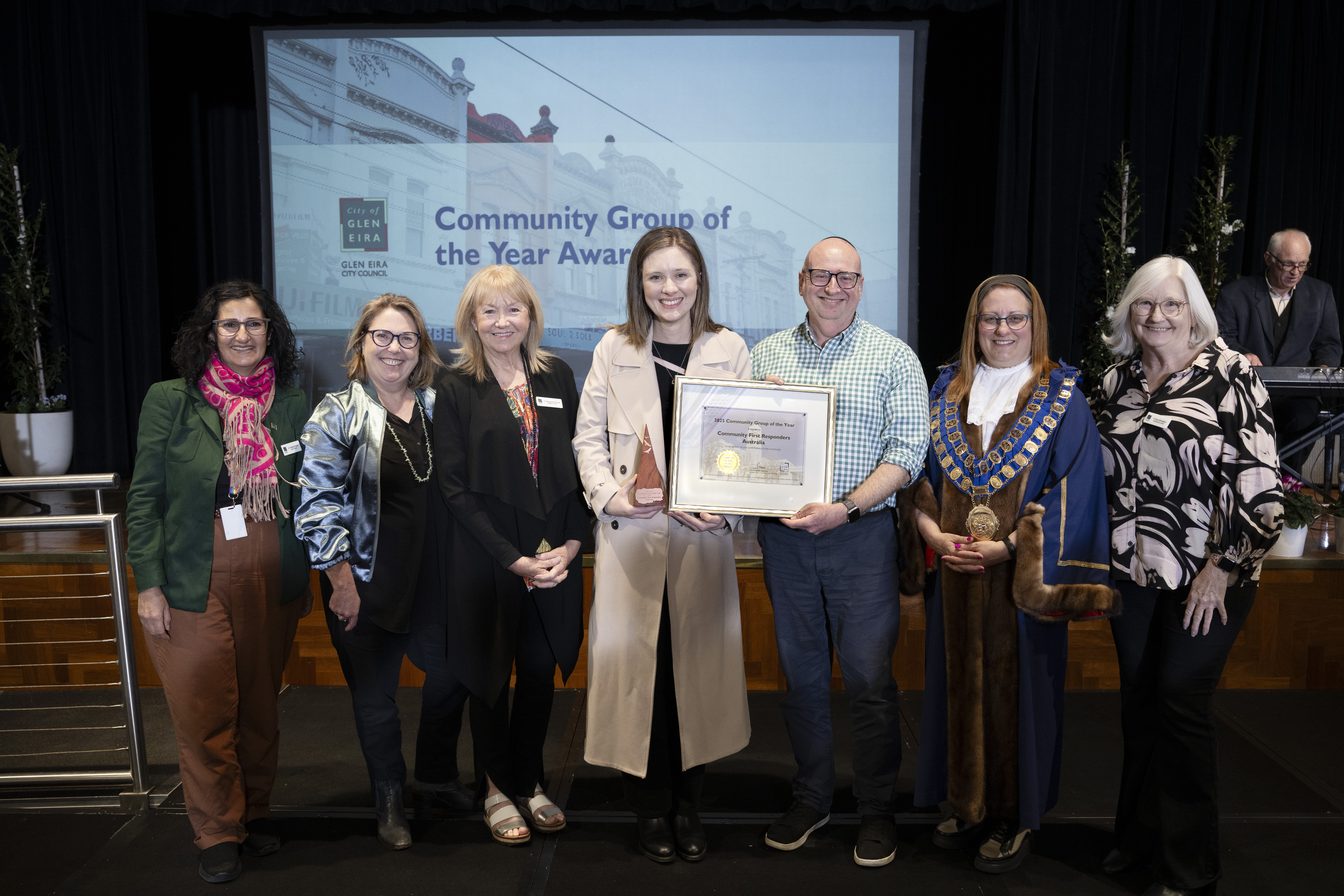 A group of people standing together on a stage, with one person in the centre holding a large framed award certificate. A projection behind them reads “Community Group of the Year Award” alongside the City of Glen Eira branding.