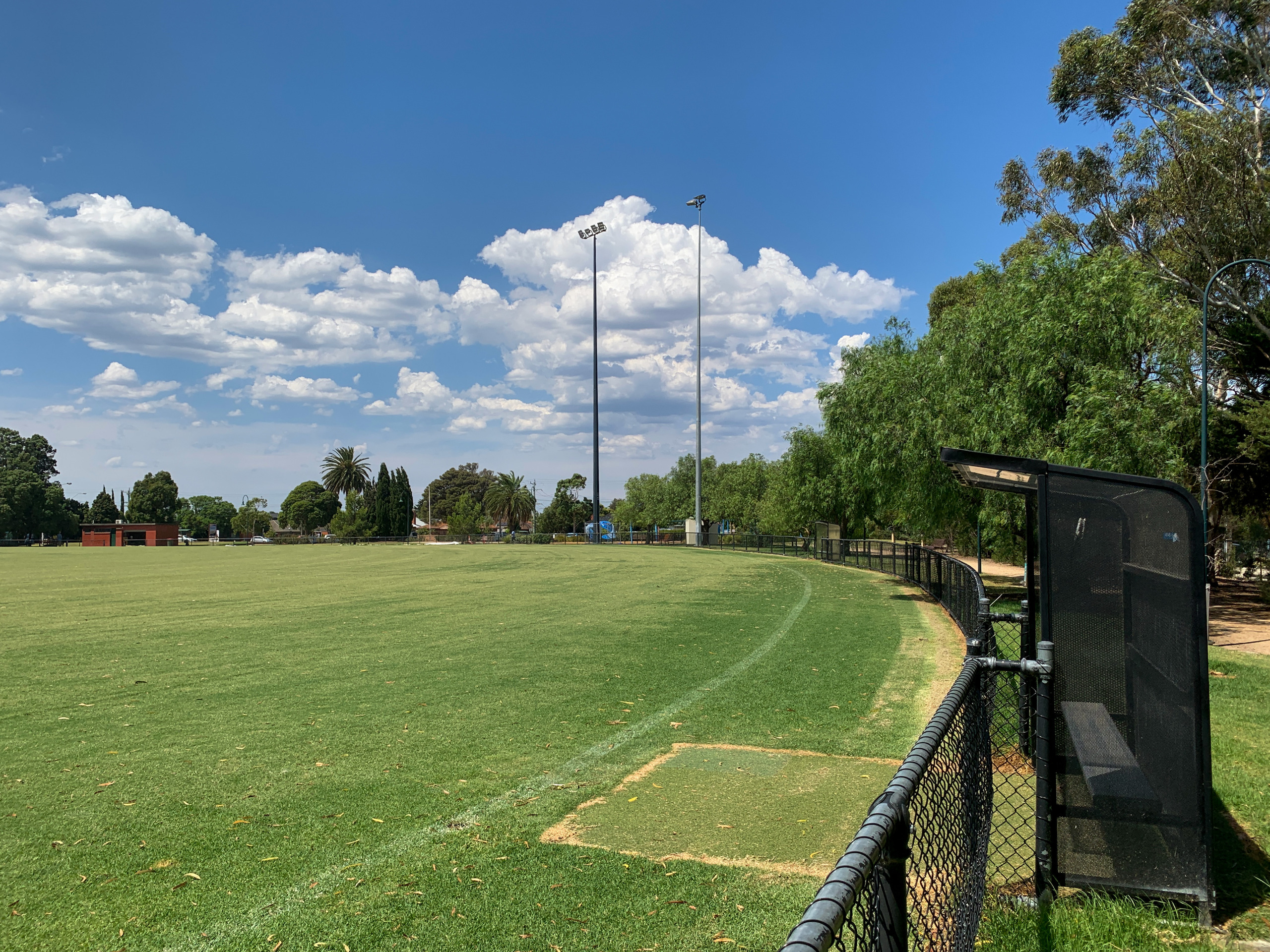 Open grass sports field bordered by a curved fence, with tall light poles and trees under a blue sky with scattered clouds.