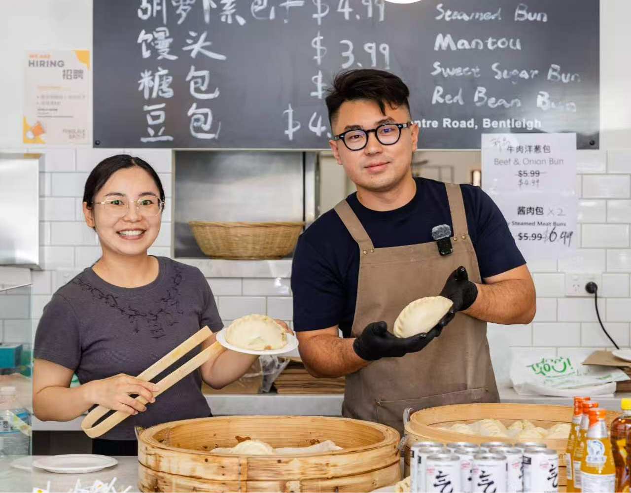 Two people standing behind a counter at a steamed bun shop, holding freshly made buns, with bamboo steamers and a menu board in the background.