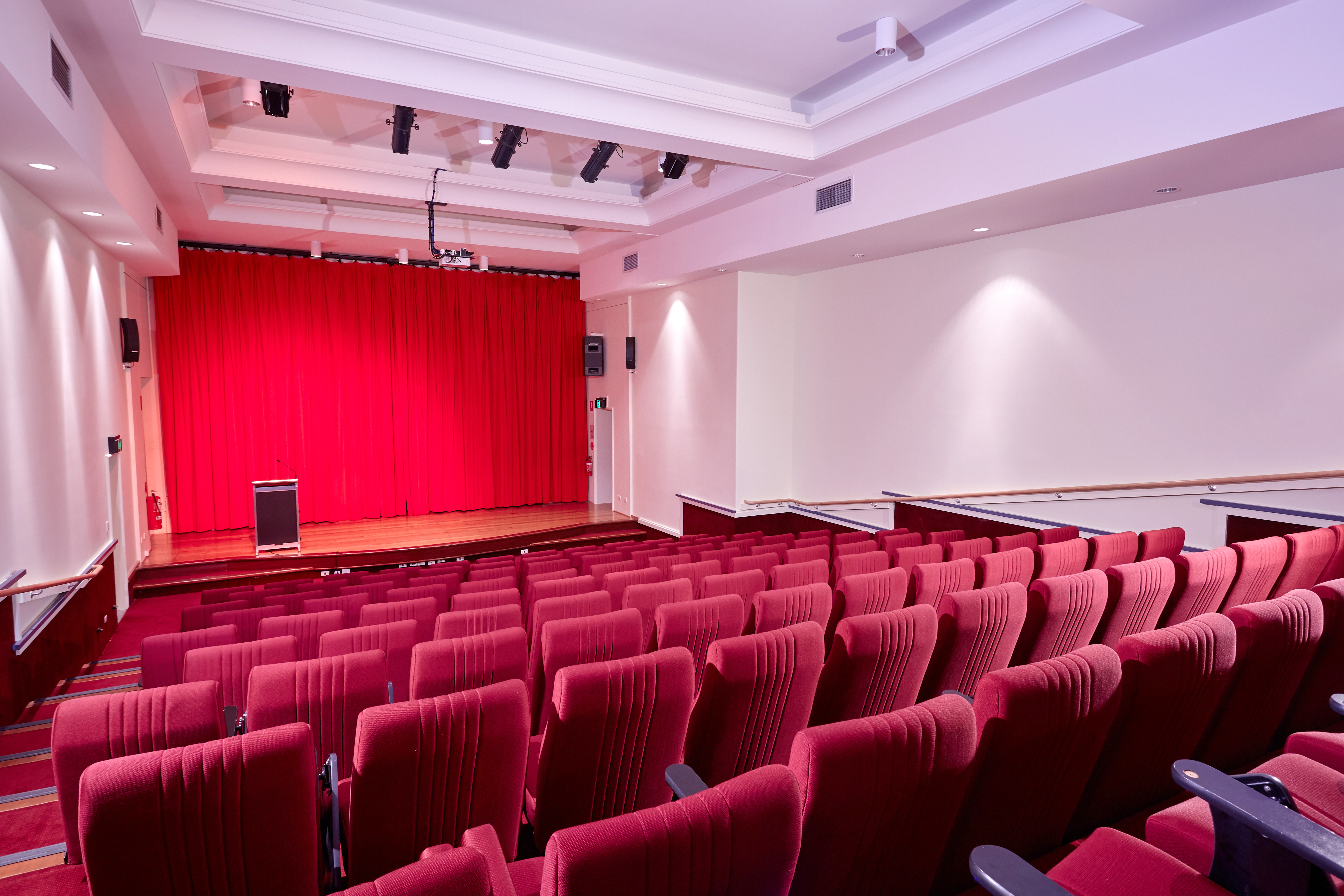 Cinema-style theatre with rows of red seats facing a stage with closed red curtains, prepared for a movie screening.
