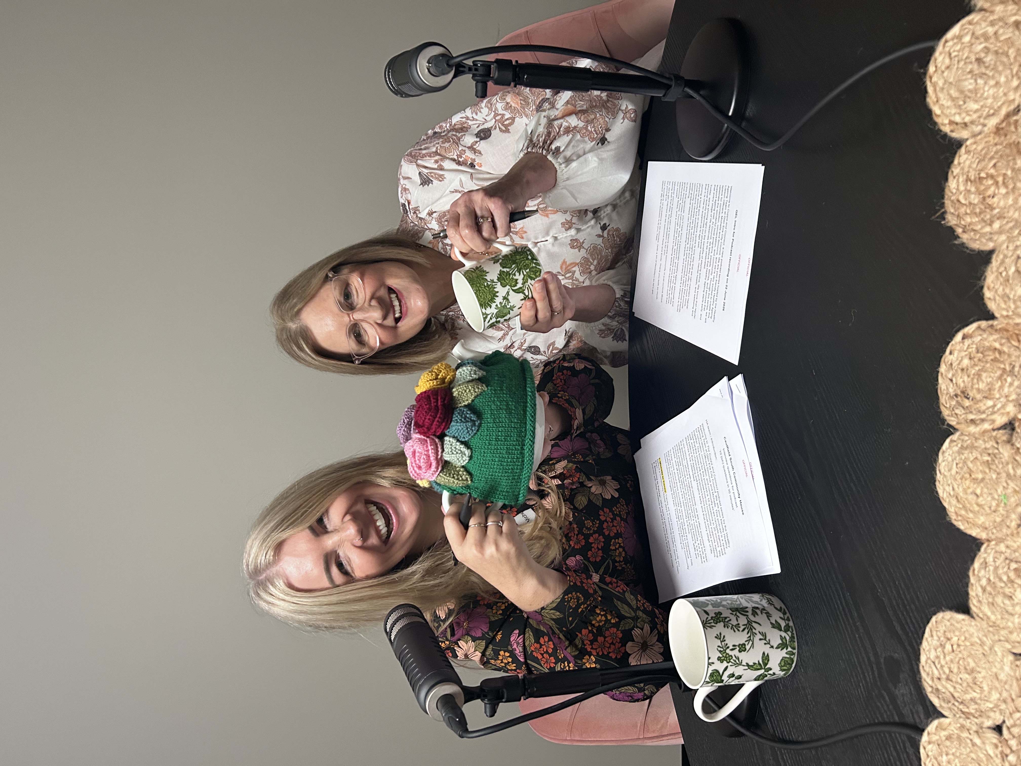 Two podcast hosts sit behind microphones holding mugs during a podcast recording session.