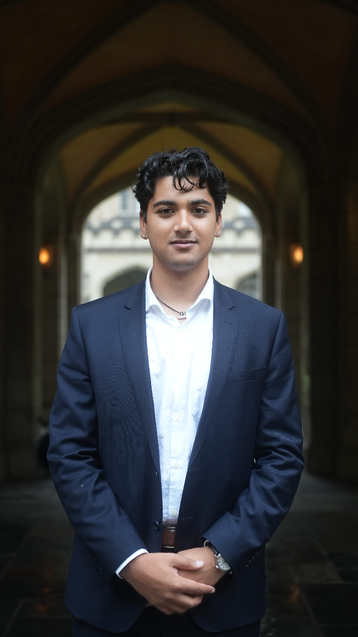 A person wearing a navy suit and white shirt standing with hands clasped in an arched stone walkway. Soft lighting highlights the architectural details in the background.