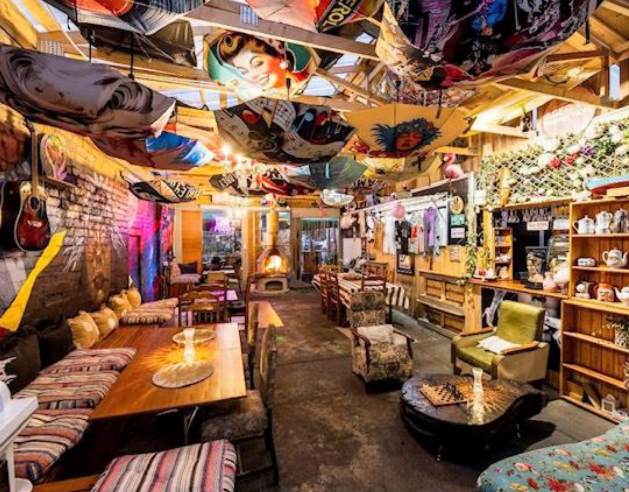 Eclectic lounge and boutique interior featuring colourful furnishings, timber tables, vintage armchairs and shelves filled with teapots and decorative items. Surfboards and artwork are suspended from the ceiling, while guitars hang on exposed brick walls. Warm lighting and a wood-fired heater create a welcoming atmosphere throughout the long, open space.