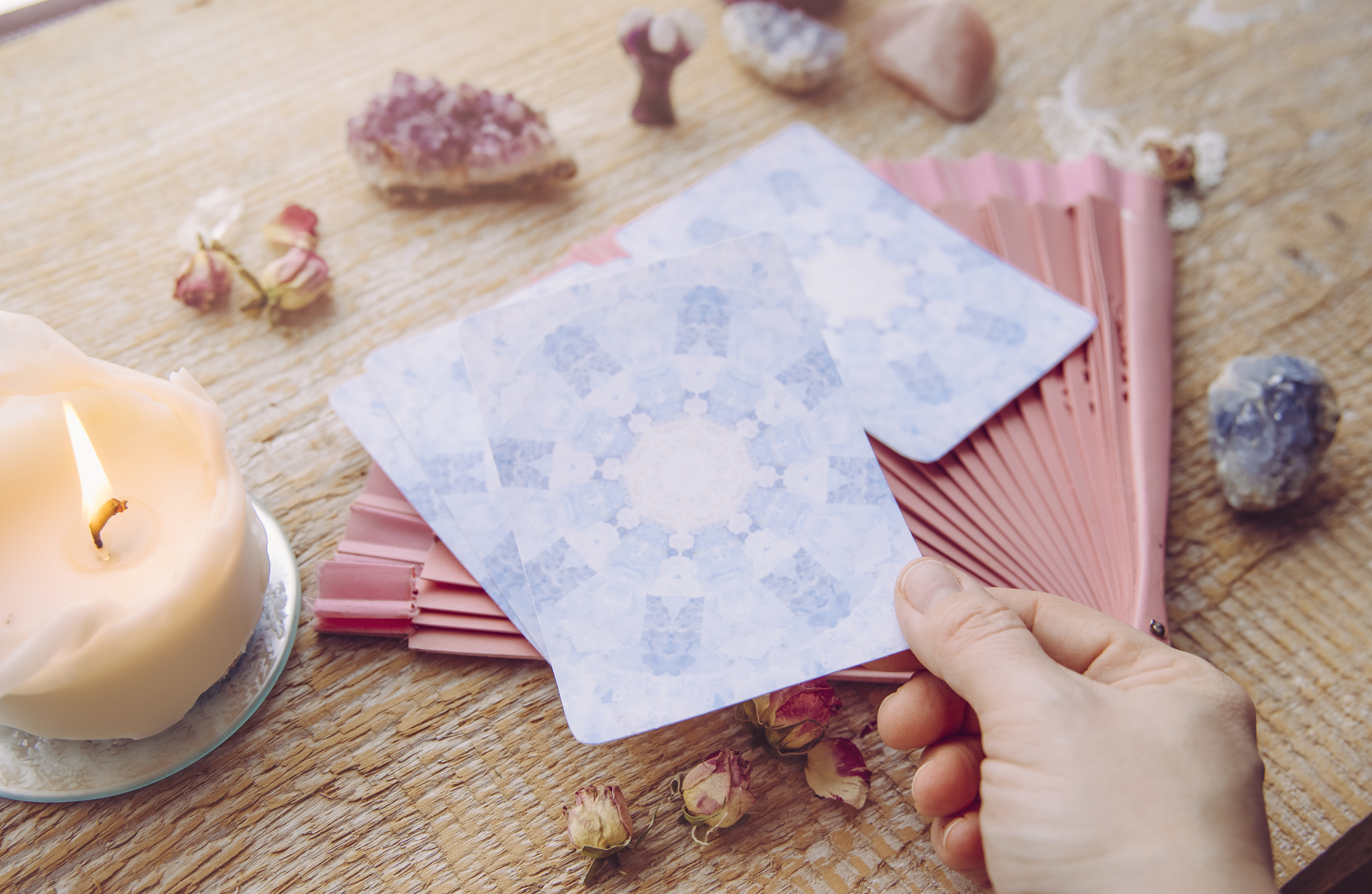 A person holds oracle cards over a table decorated with crystals, dried flowers and a candle during a creative oracle card-making activity.
